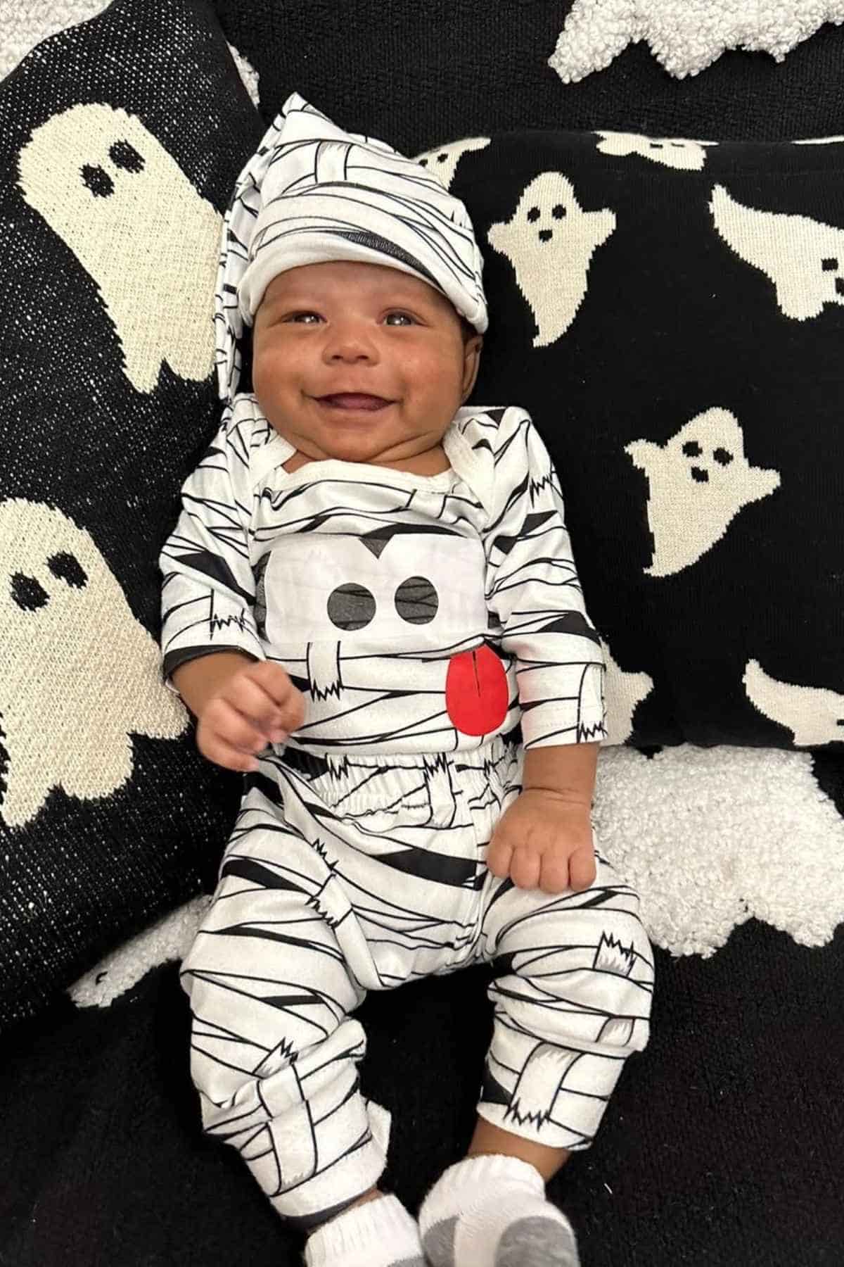 A baby dressed with a patterned black and white onesie paired with hat while lying on a ghost-patterned sheet.