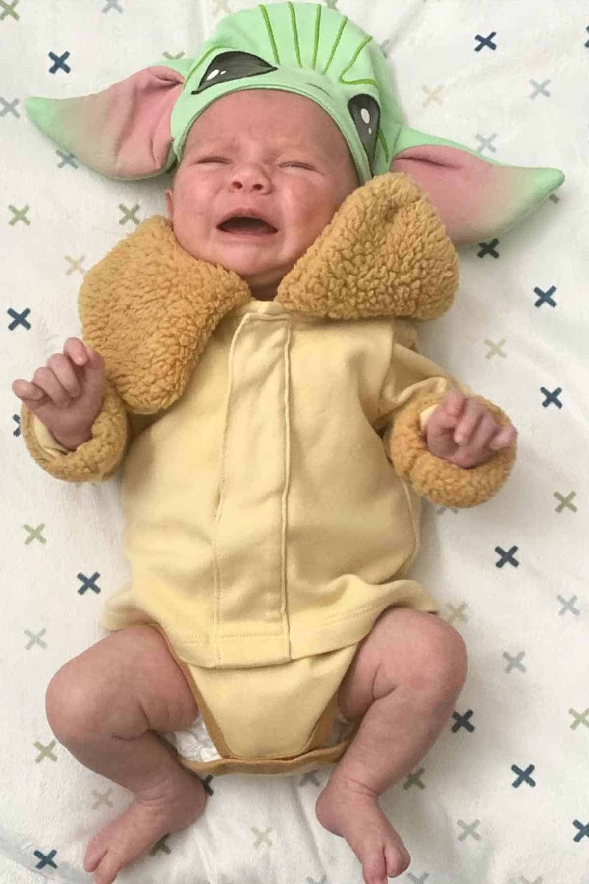 A baby wearing a yellowish onesie and a grugo headpiece while lying on a pattern sheet.