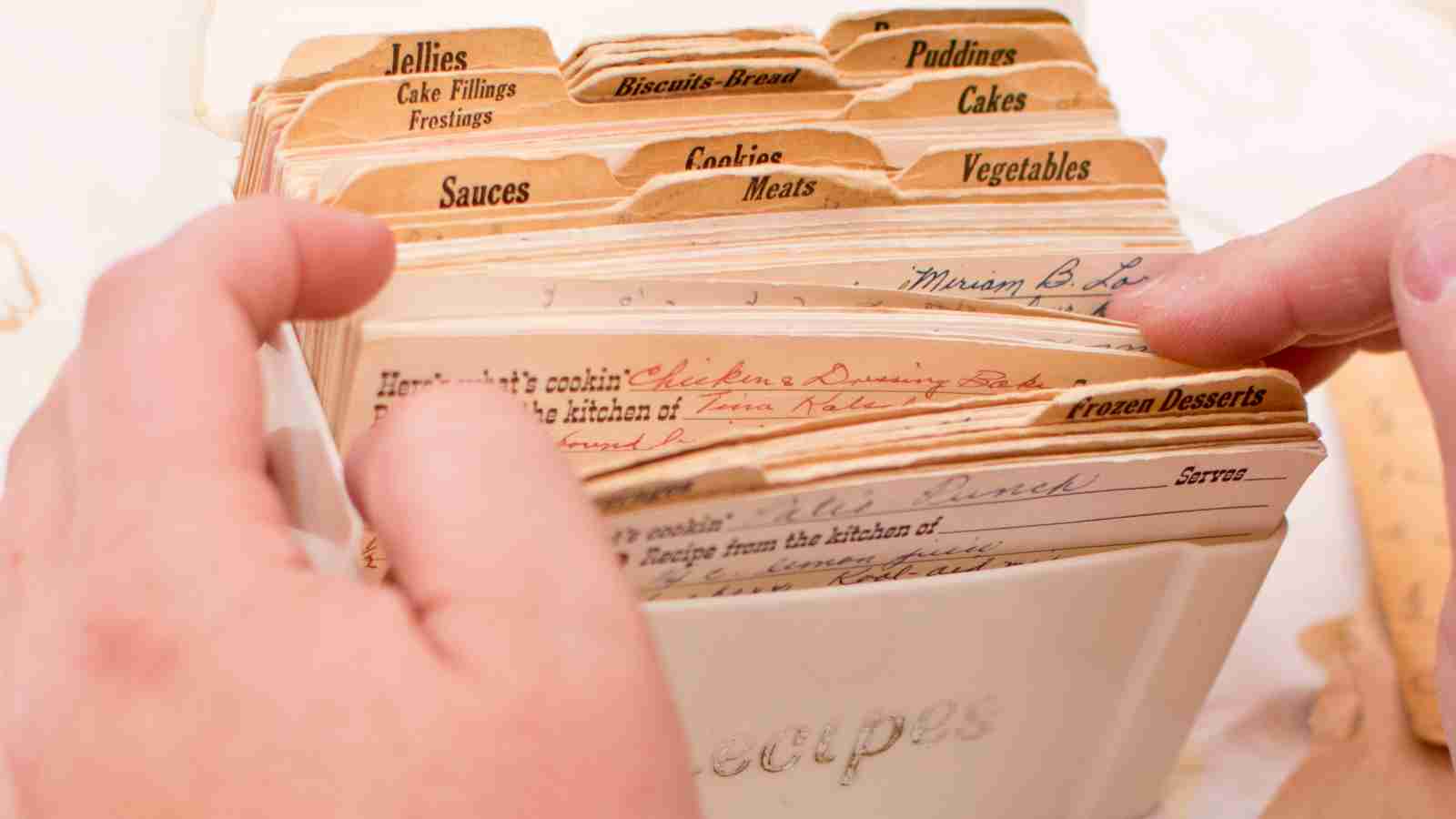 A person's hands are shown looking through a vintage recipe box filled with recipe cards.