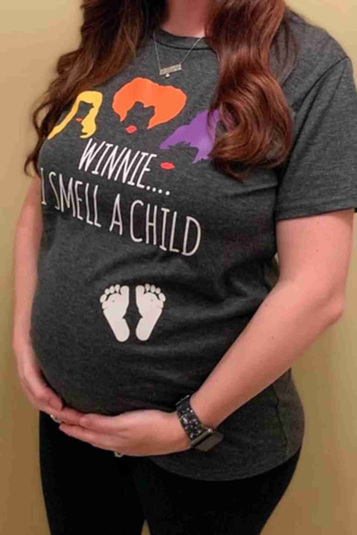 A pregnant woman wearing a T-shirt with illustrations of three heads, the text "Winnie... I smell a child," and an image of baby footprints over her belly.
