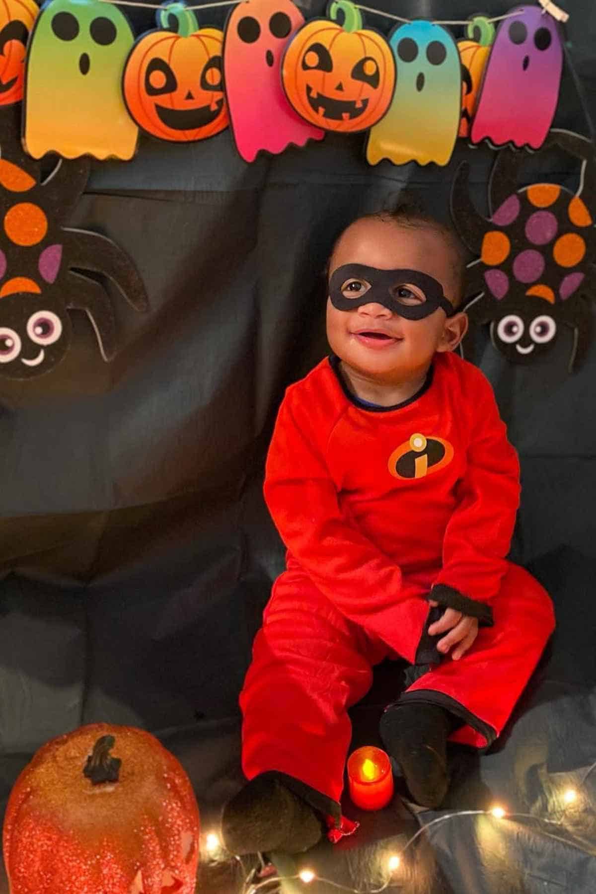 A young child in an Incredibles costume sits on a black backdrop decorated with Halloween-themed pumpkins, ghosts, spiders, string lights, and a pumpkin prop.