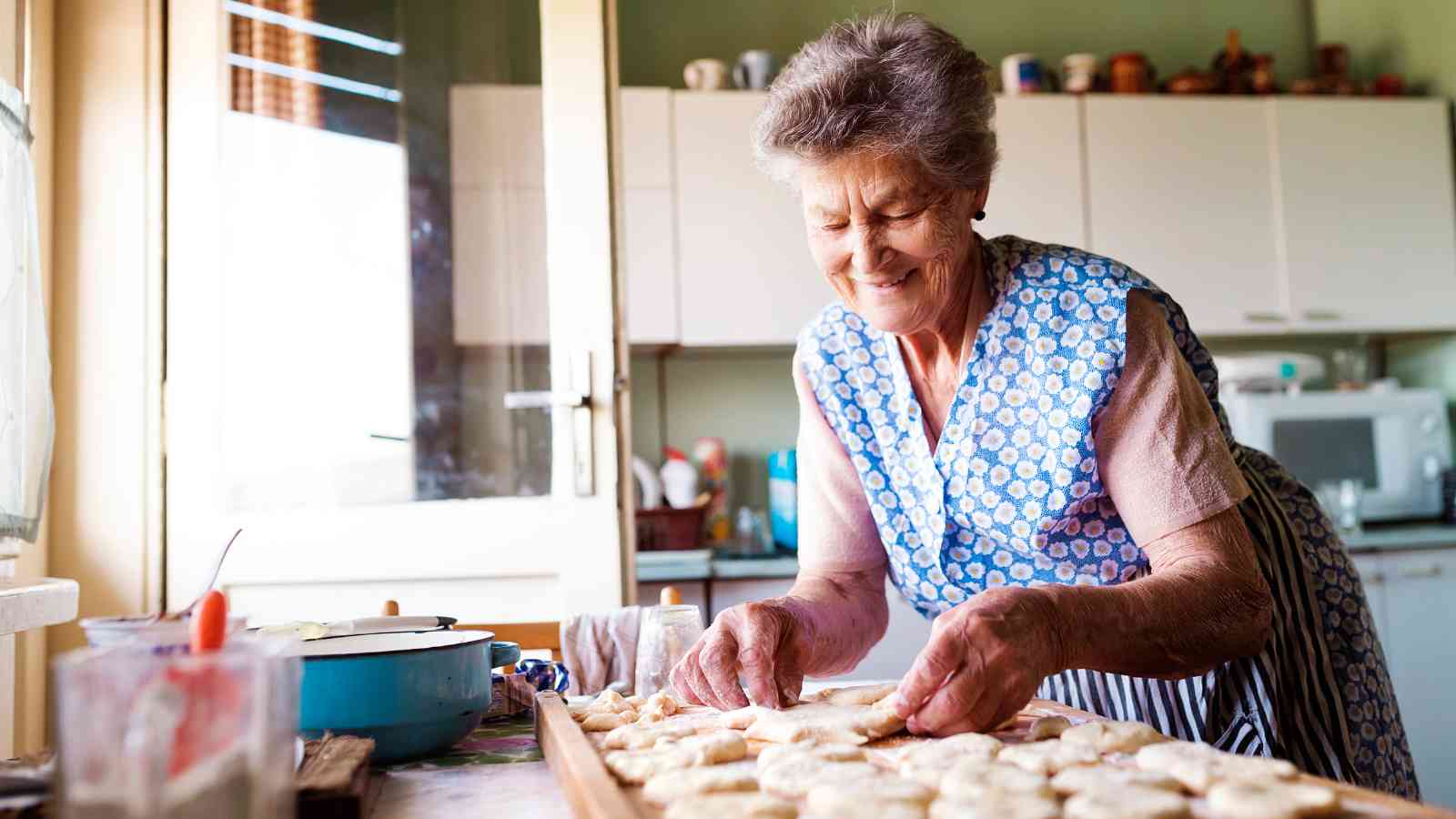 An elderly woman smiles as she prepares dough for baking in her kitchen.