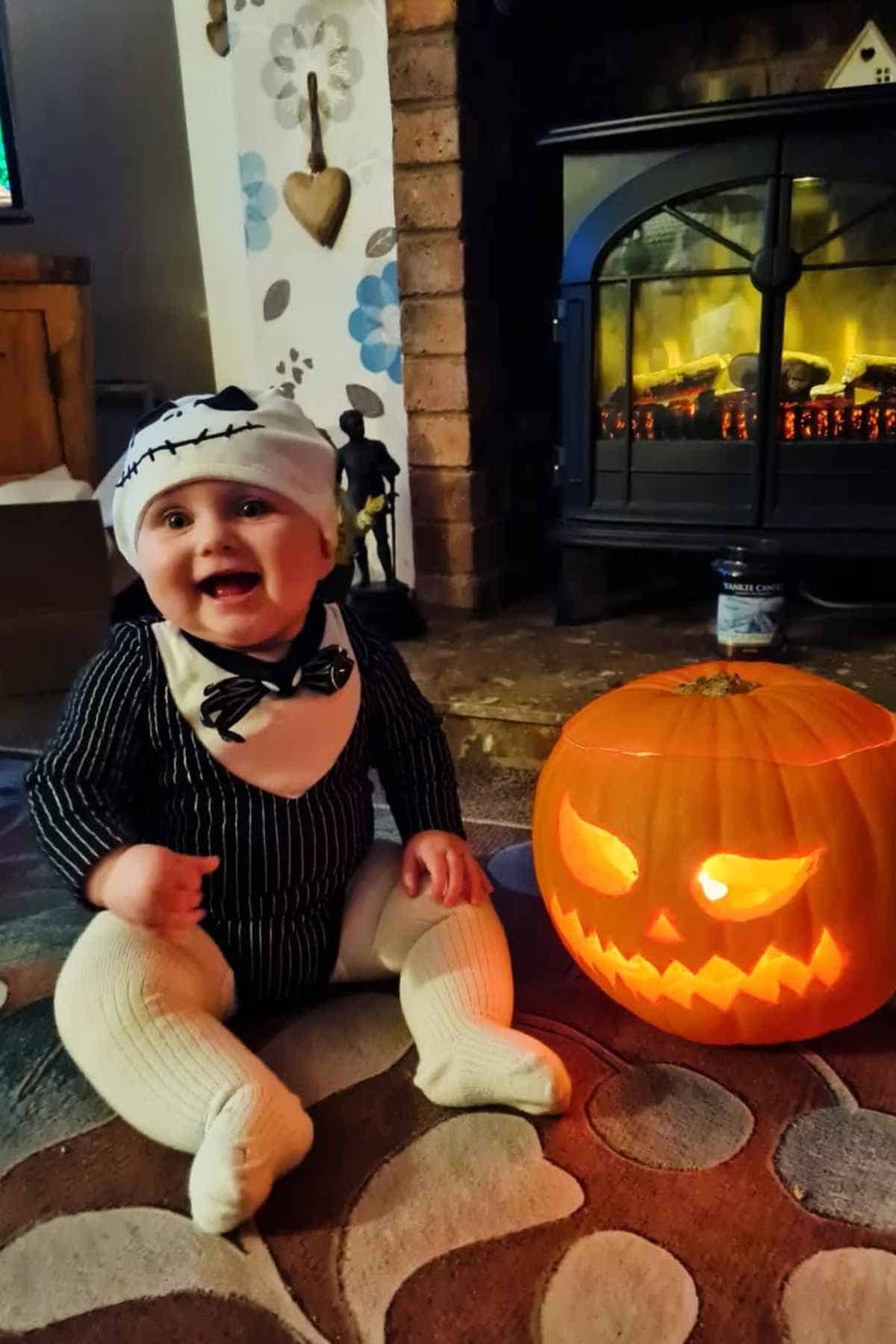 A baby dressed in a black and white costume sits on a rug next to a carved, lit jack-o’-lantern in front of a fireplace.