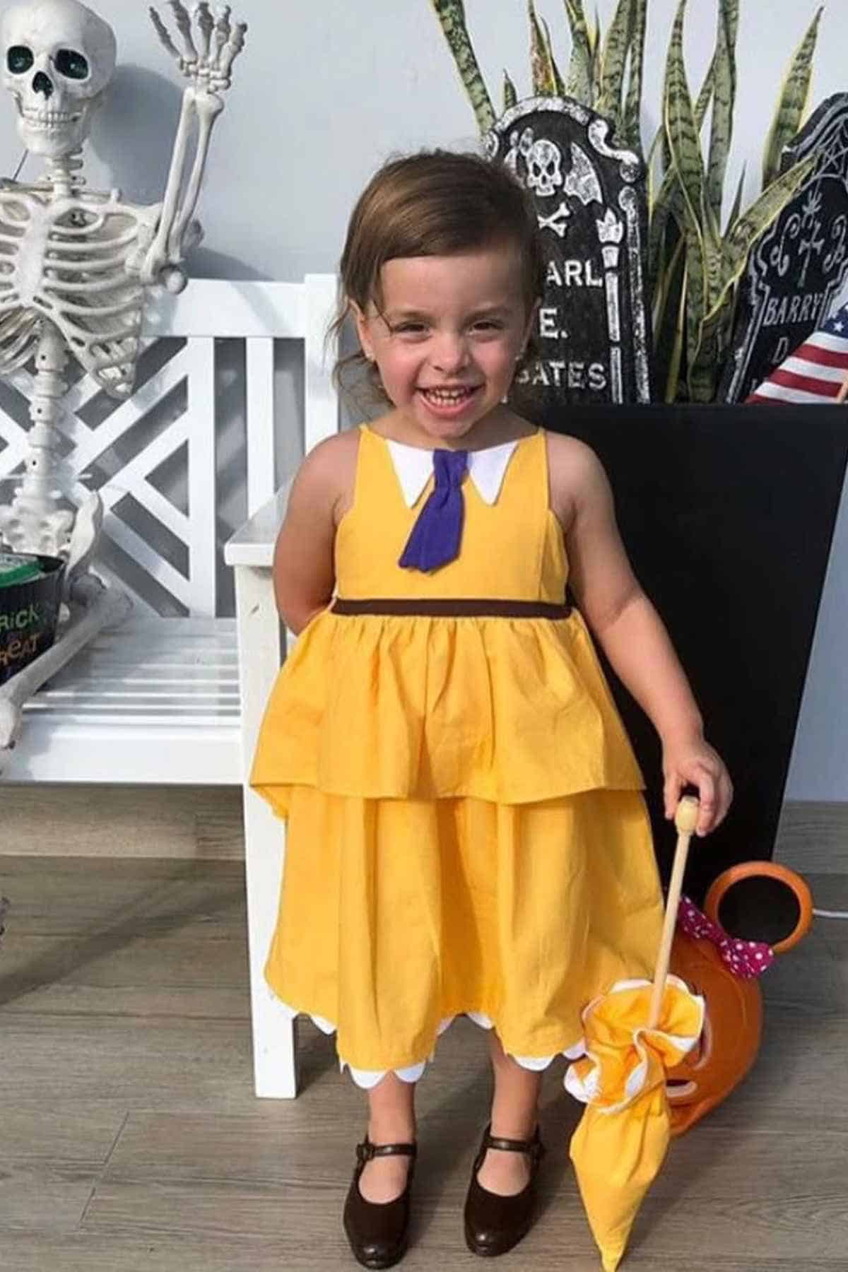 A young child in a yellow layered dress with a blue tie stands indoors, smiling, holding an umbrella, with Halloween decorations in the background.