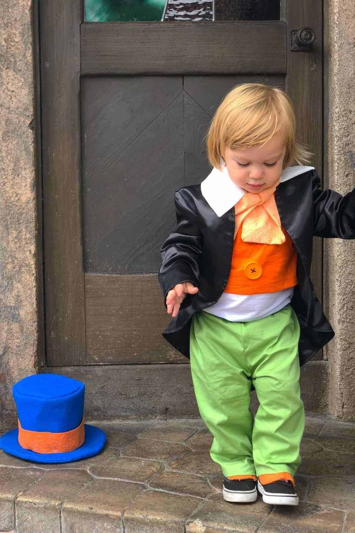 A toddler dressed in a colorful costume with a black jacket, orange vest, green pants, and an oversized collar stands next to a blue and orange top hat on the ground in front of a door.