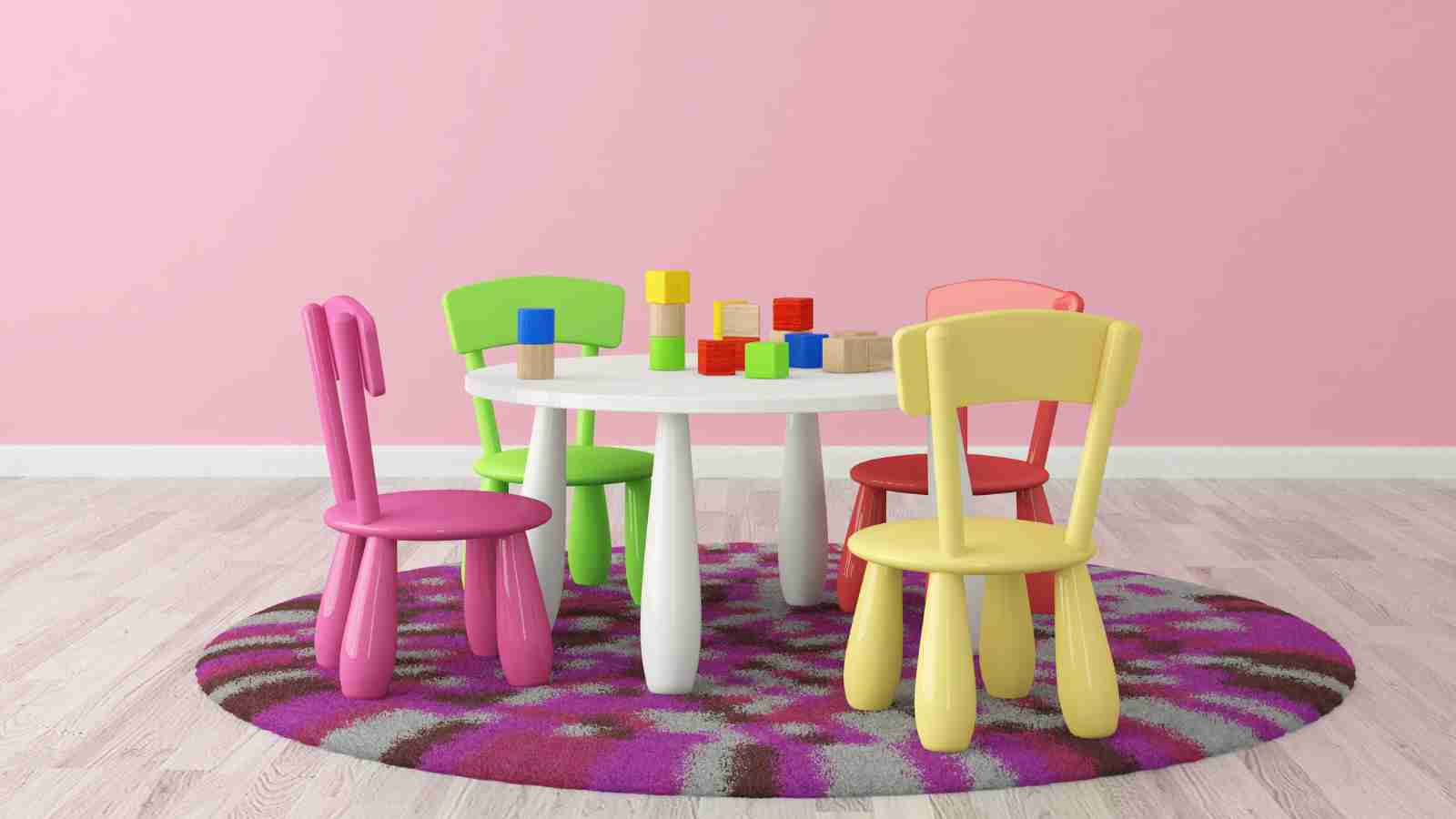 A round white table with colorful building blocks and four brightly colored chairs stands on a purple patterned rug against a pink wall in a children's playroom.