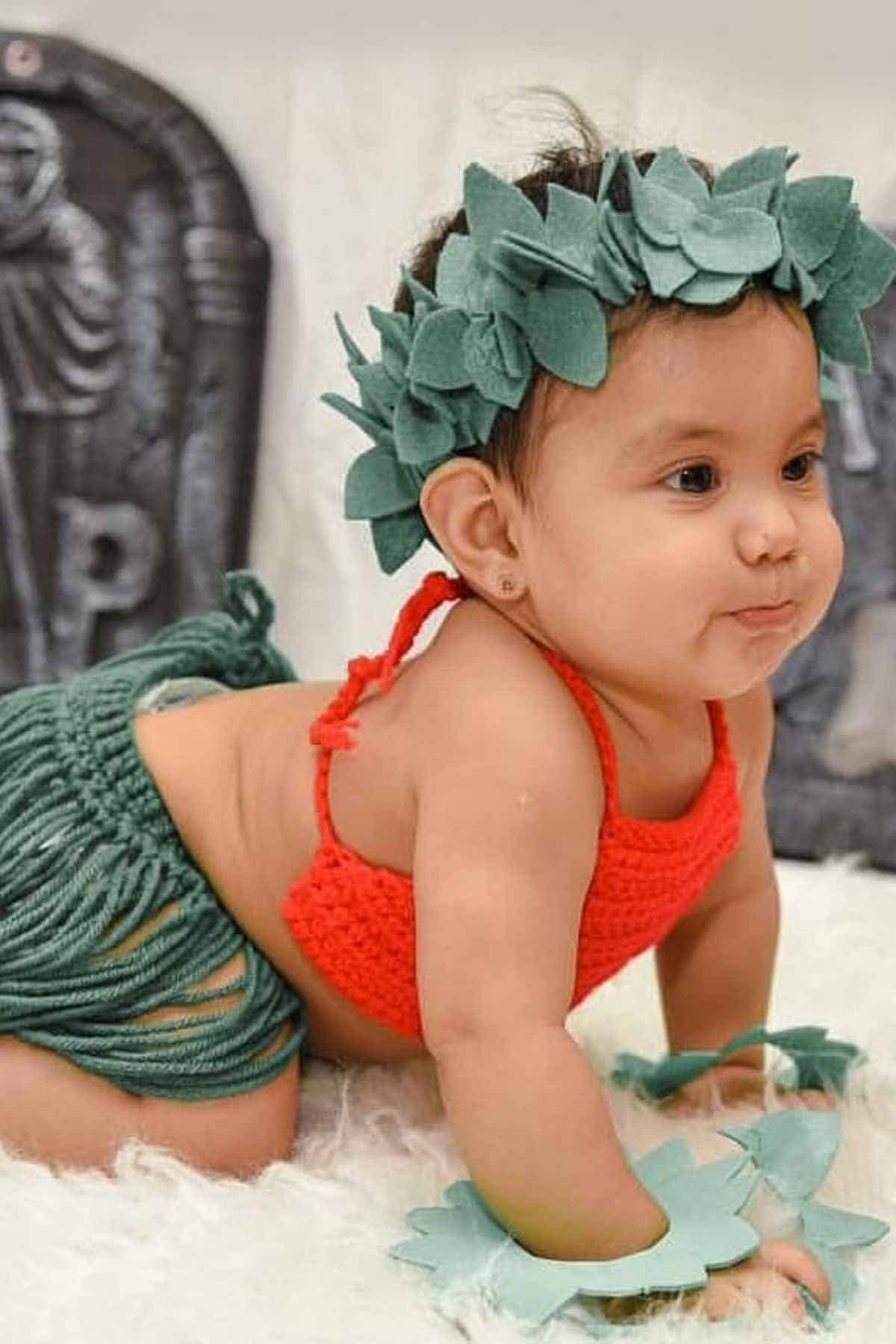 A baby wearing a red crochet top, green grass-style skirt, leaf crown, and bracelets crawls on a white surface with gray decorations in the background.
