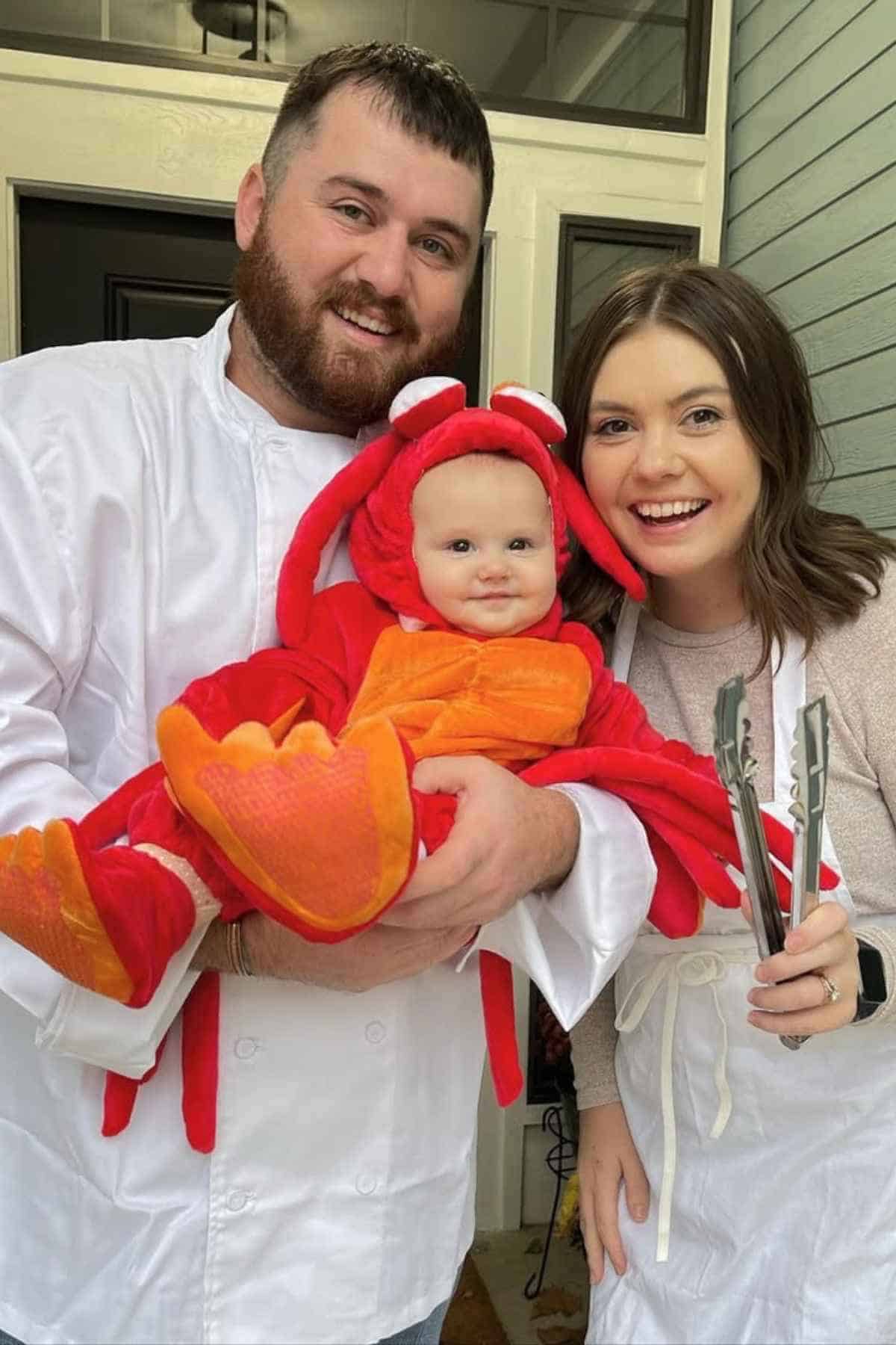 Two adults stand smiling, one holding a baby dressed in a red lobster costume. The adults wear white kitchen attire; one holds metal tongs. They pose in front of a door.