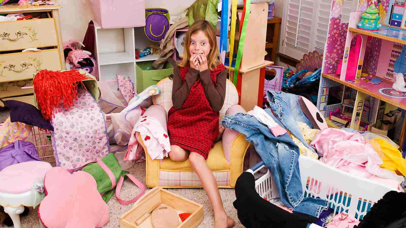 A young girl sits amidst a very messy room, looking overwhelmed by the clutter of toys and clothes.