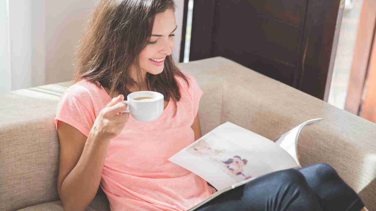 A smiling woman sits on a couch, relaxing with a cup of coffee and a magazine.