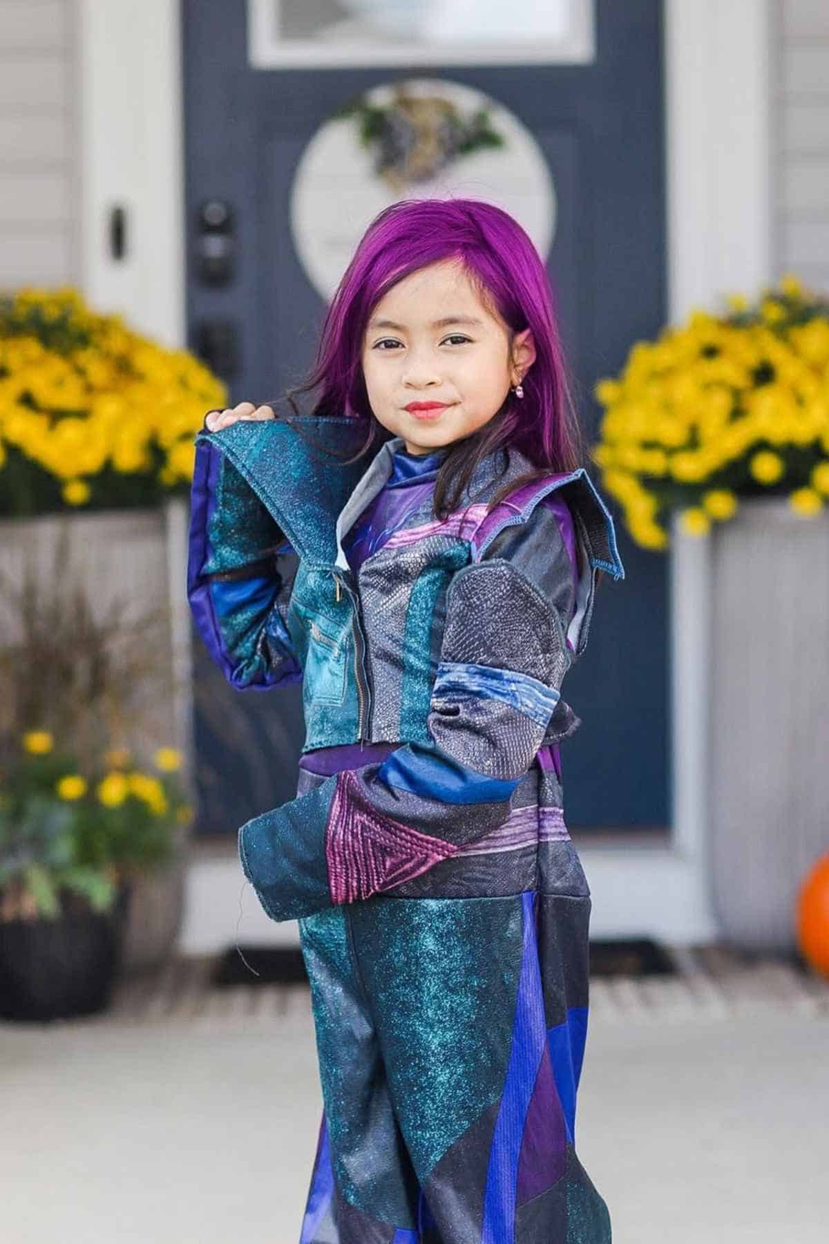A young girl with purple hair poses in a shiny, multi-colored costume in front of a door with yellow flowers on either side.