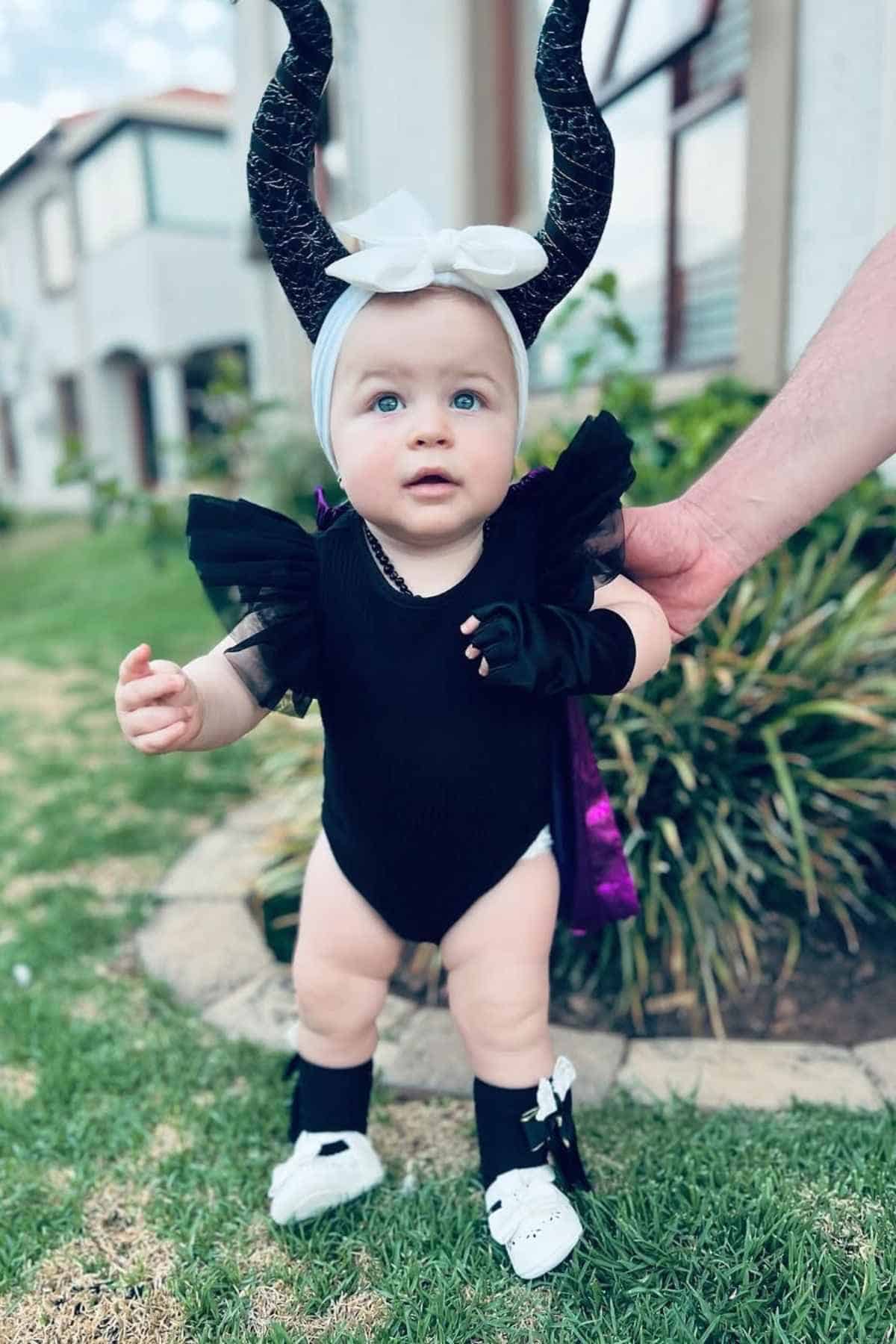 A baby dressed in a black Maleficent costume with horned headband stands outside on grass, assisted by an adult's hand.