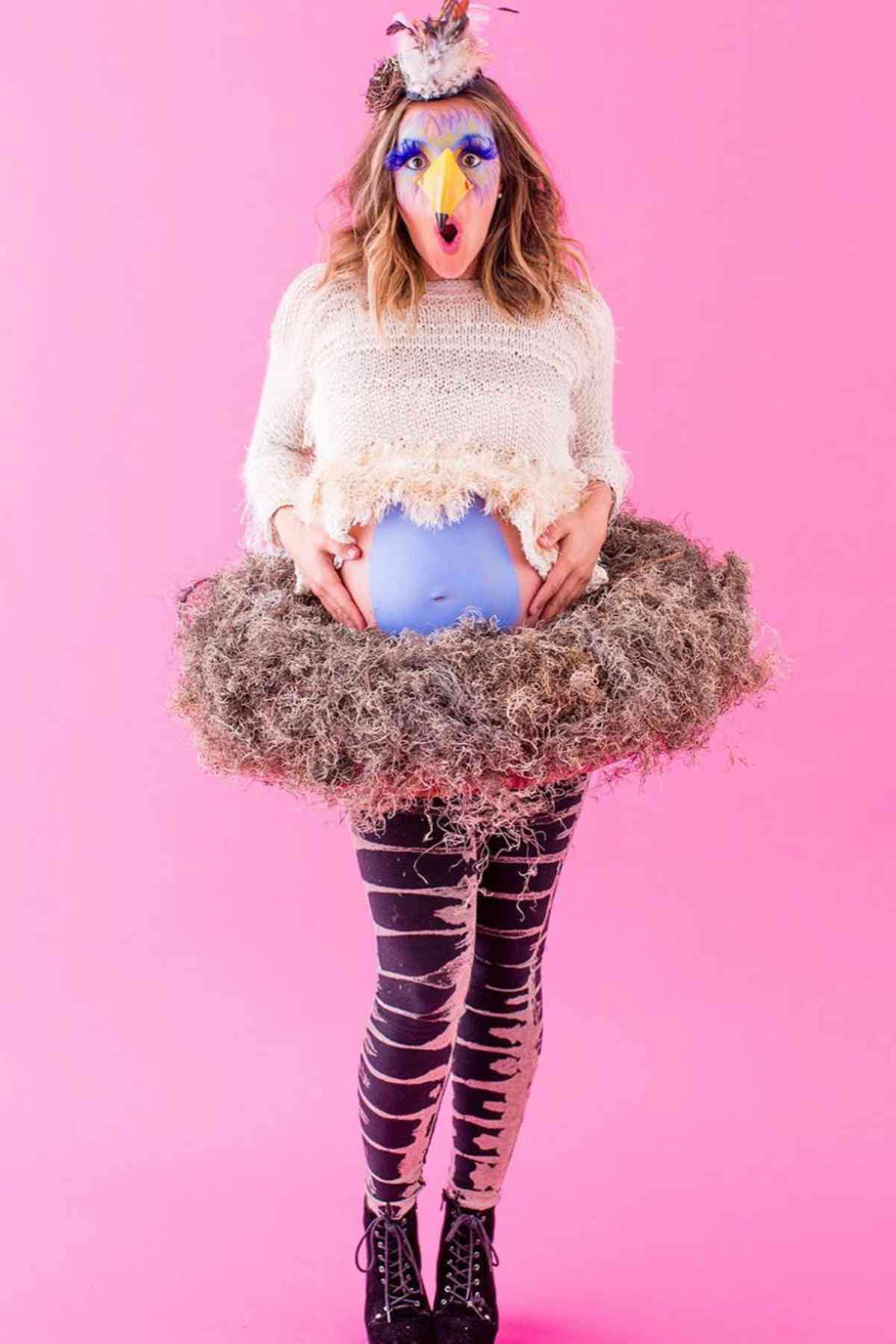 A person wearing a bird mask, a white sweater, nest-like skirt, striped leggings, and with a painted blue belly stands against a pink background.