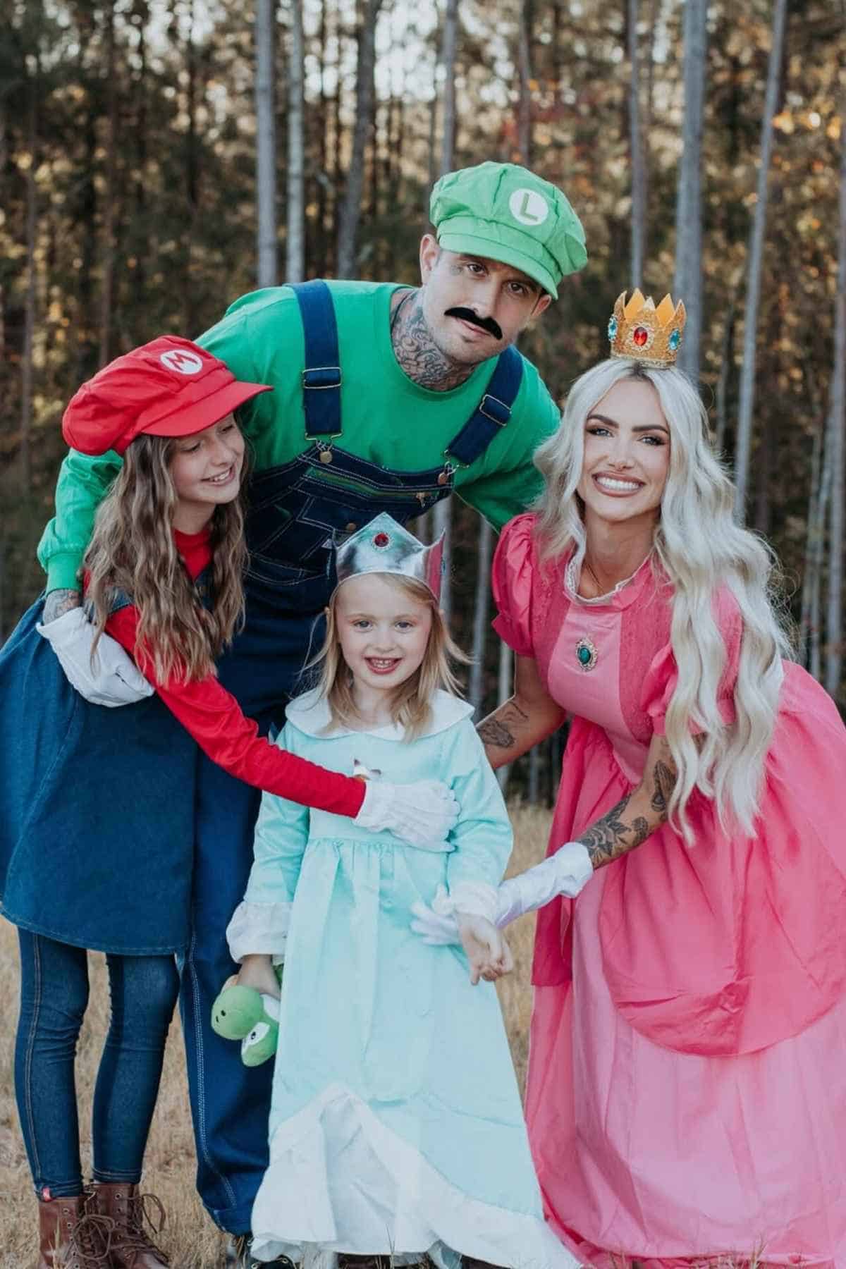 Four people dressed in Mario-themed costumes—Mario, Luigi, Princess Peach, and Rosalina—pose together outdoors in front of trees.