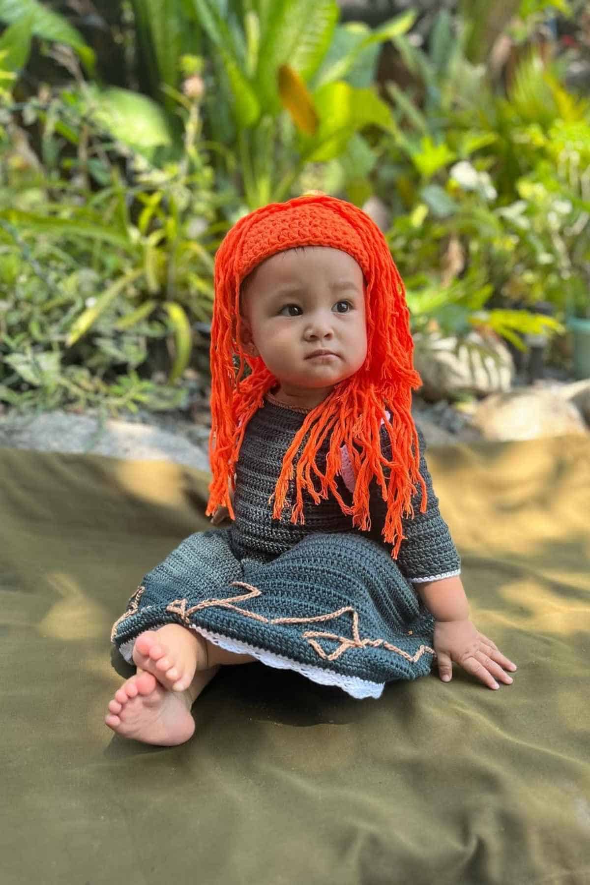 A baby sits outdoors on a green blanket, wearing a dark knit dress and an orange crochet wig, with plants in the background.