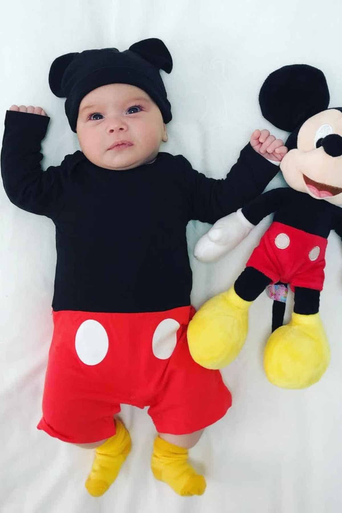A baby dressed in a Mickey Mouse costume lies on a white surface next to a Mickey Mouse stuffed toy.