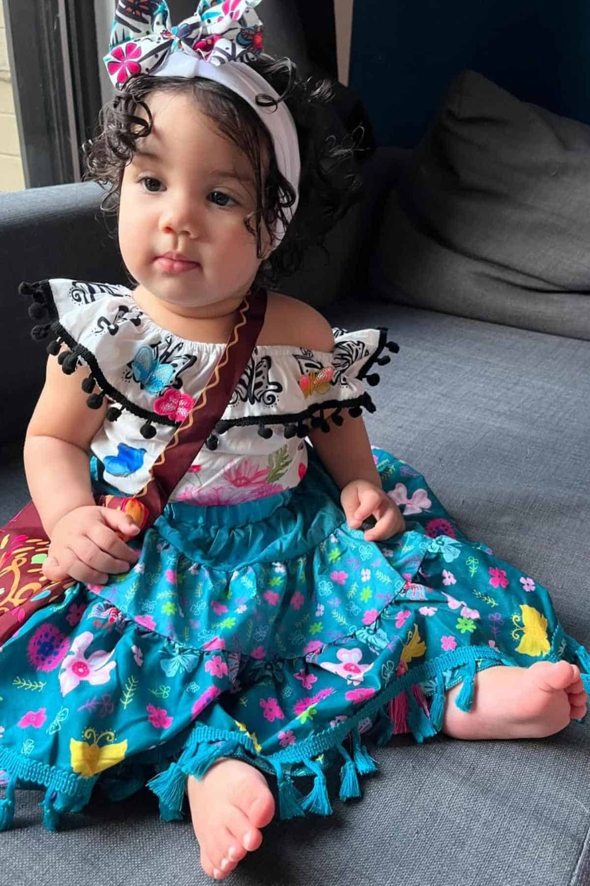 A young girl with curly hair sits on a gray couch, wearing a colorful dress and a matching headband.