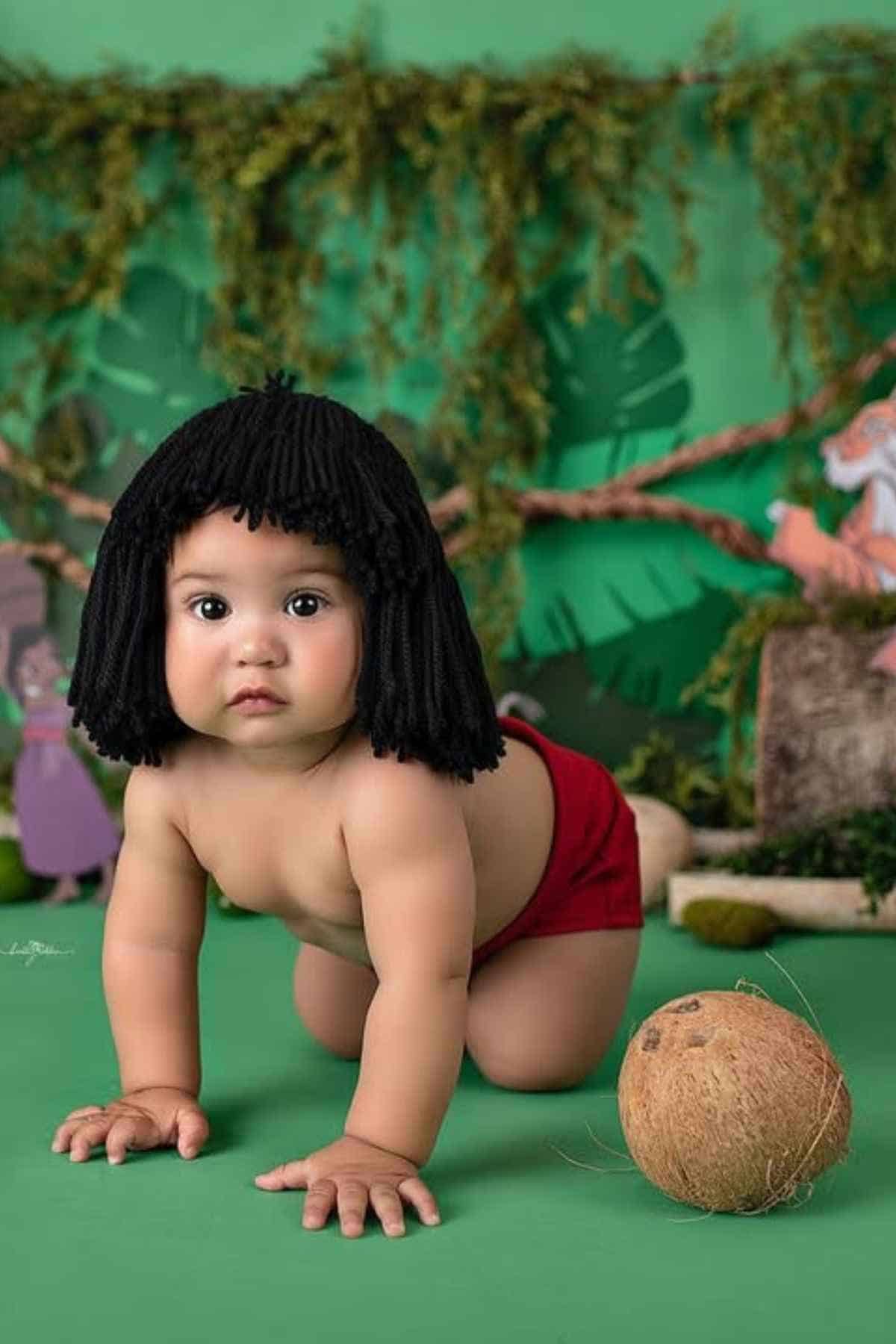 A baby wearing a black wig and red shorts crawls on a green surface with a coconut nearby and jungle-themed decorations in the background.