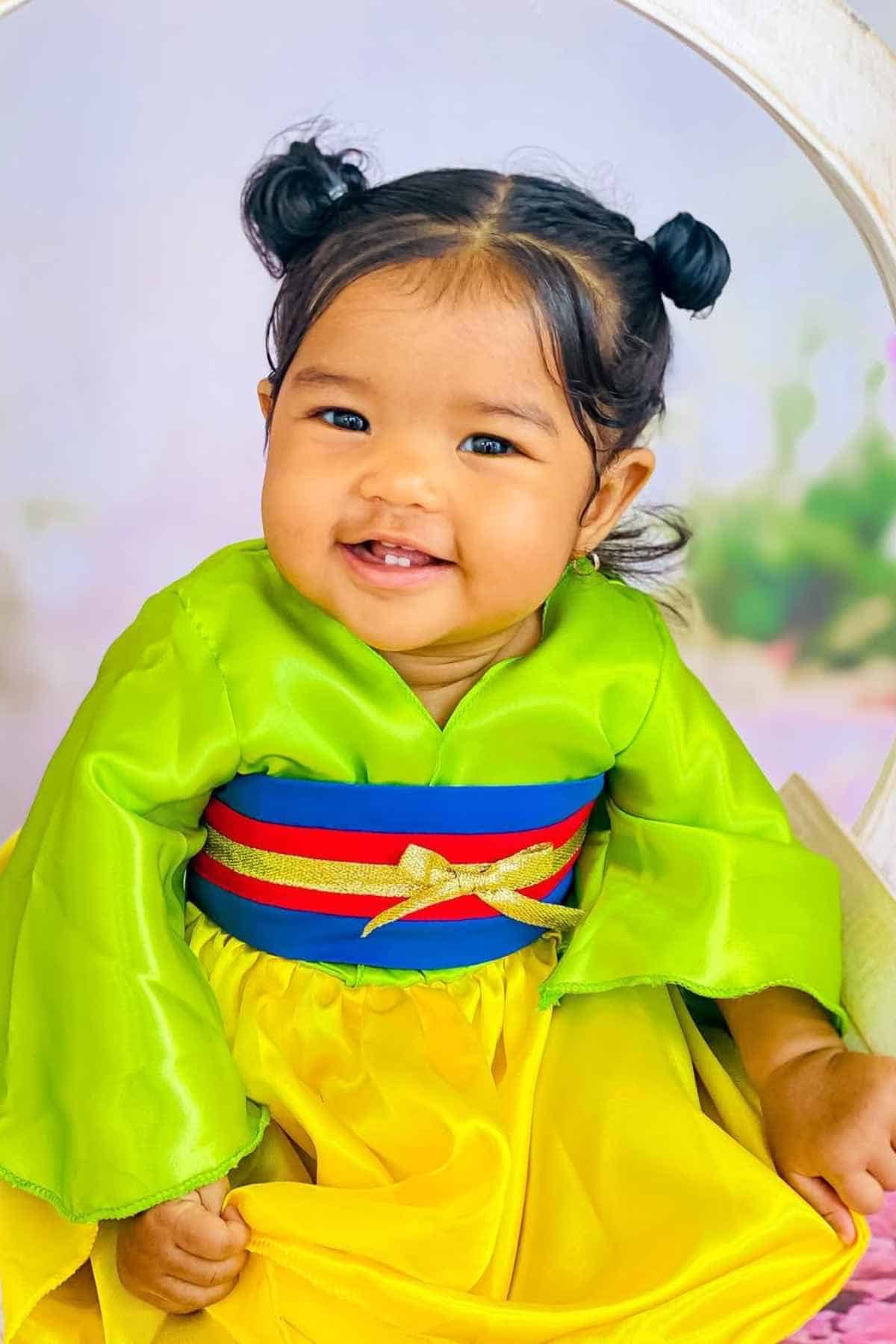 Smiling baby wearing a bright green and yellow outfit with a colorful sash, sitting and looking at the camera.