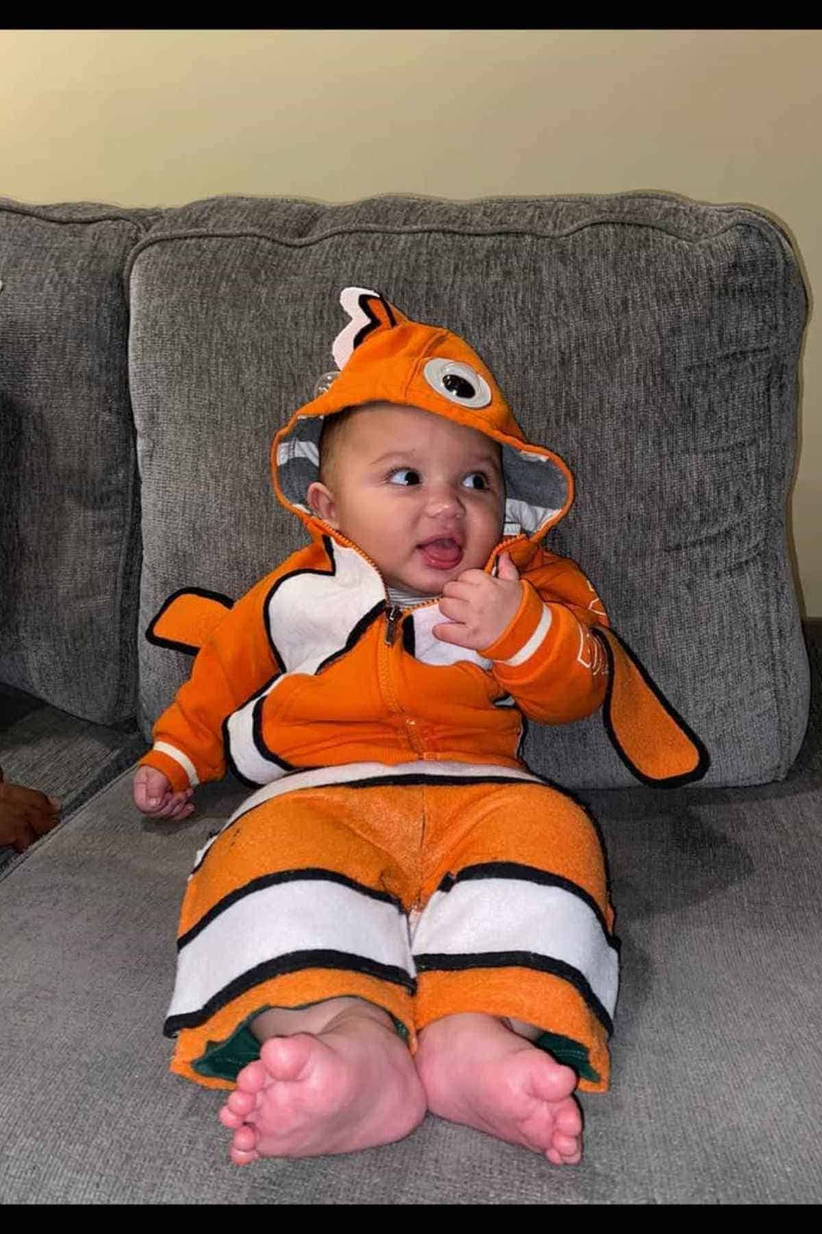 A baby sits on a gray couch wearing an orange and white clownfish costume with a hood featuring eyes and fins.