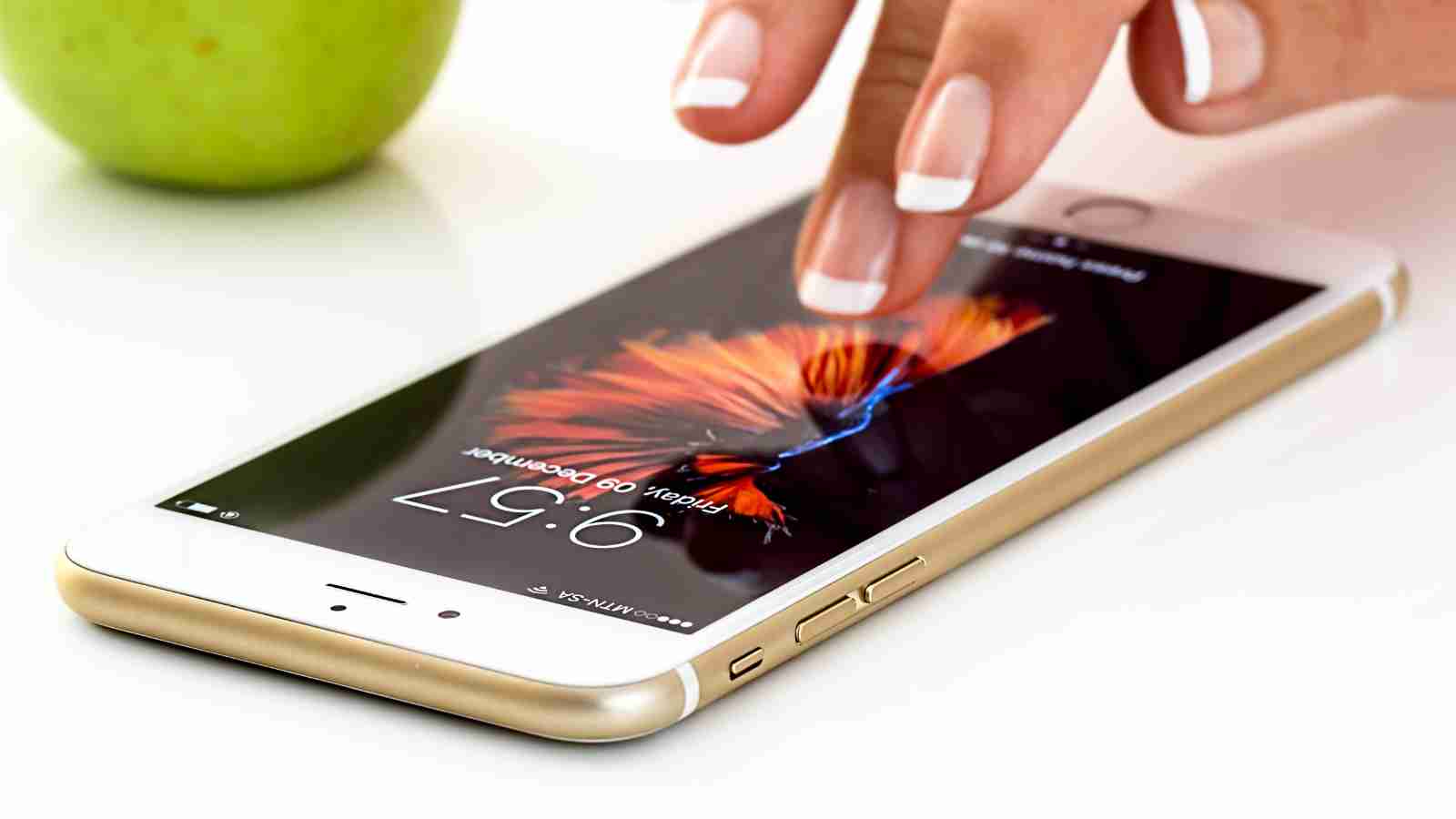 A hand with French manicured nails touches the screen of a gold smartphone displaying a fish image on its lock screen.