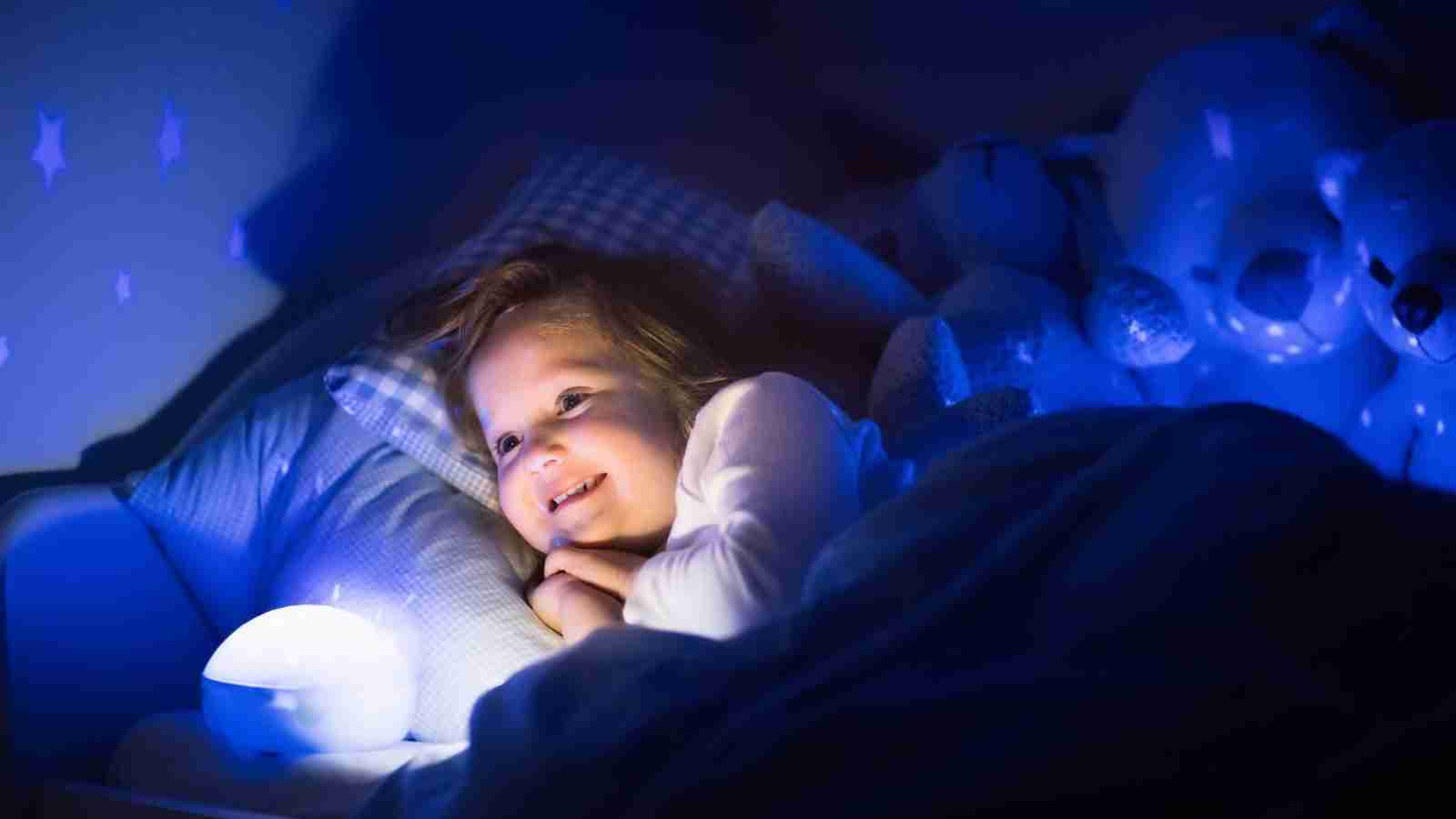 A young child lies in bed at night, smiling and illuminated by a glowing night light, with stuffed animals in the background.