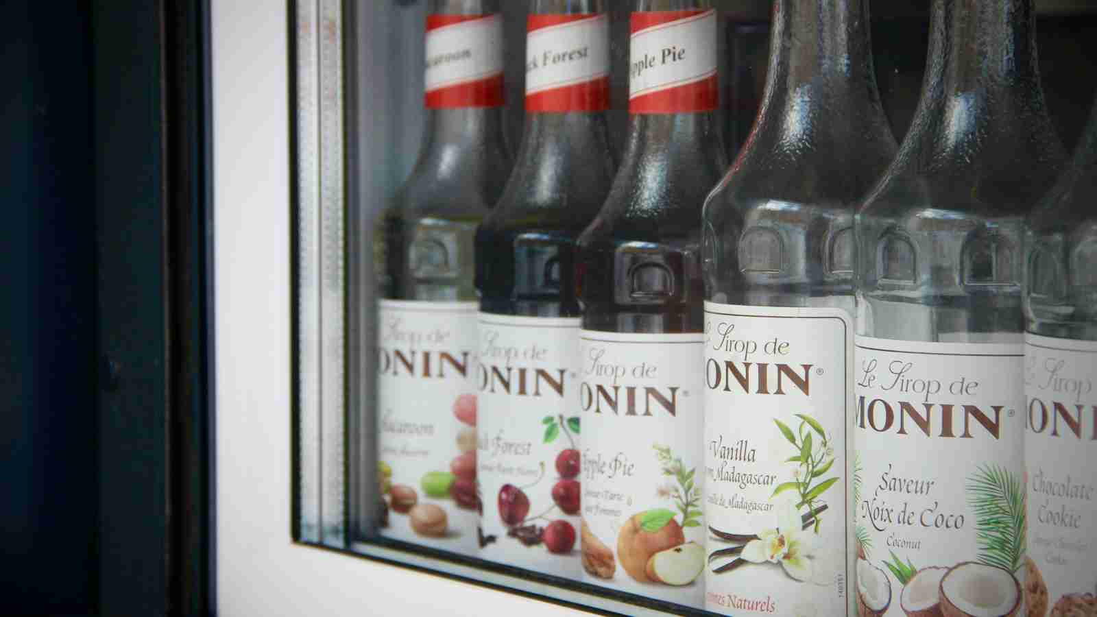 Several bottles of Monin flavored syrups are displayed in a refrigerated case.
