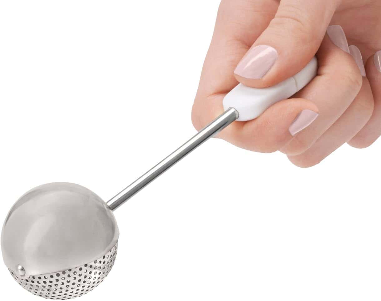 A hand holding a metal tea infuser with a white handle.