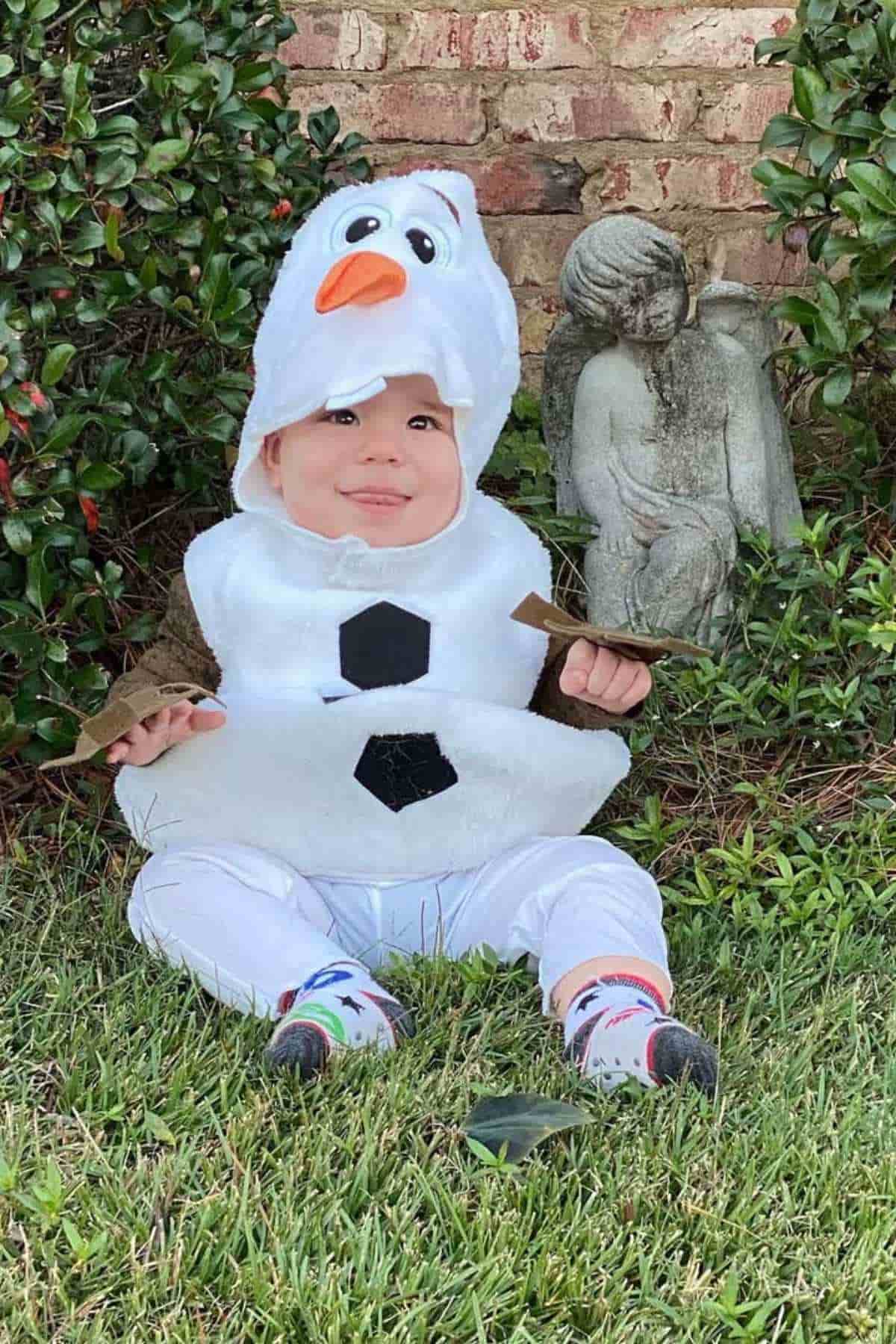 A baby sits on grass wearing a snowman costume with a hood, next to a small stone angel statue and some greenery.