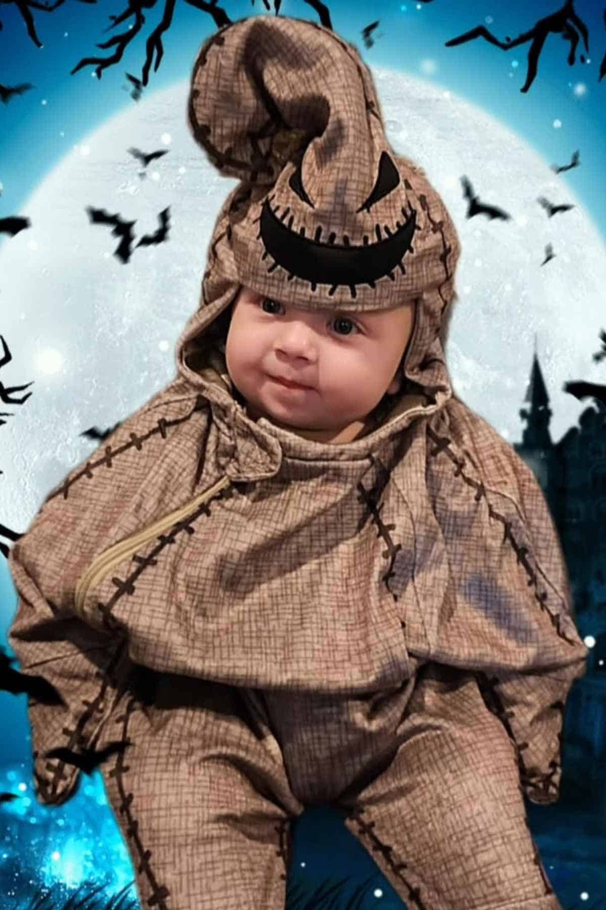 A baby dressed in a burlap costume with black stitching and a hood is sitting in front of a moonlit background with bats and trees.