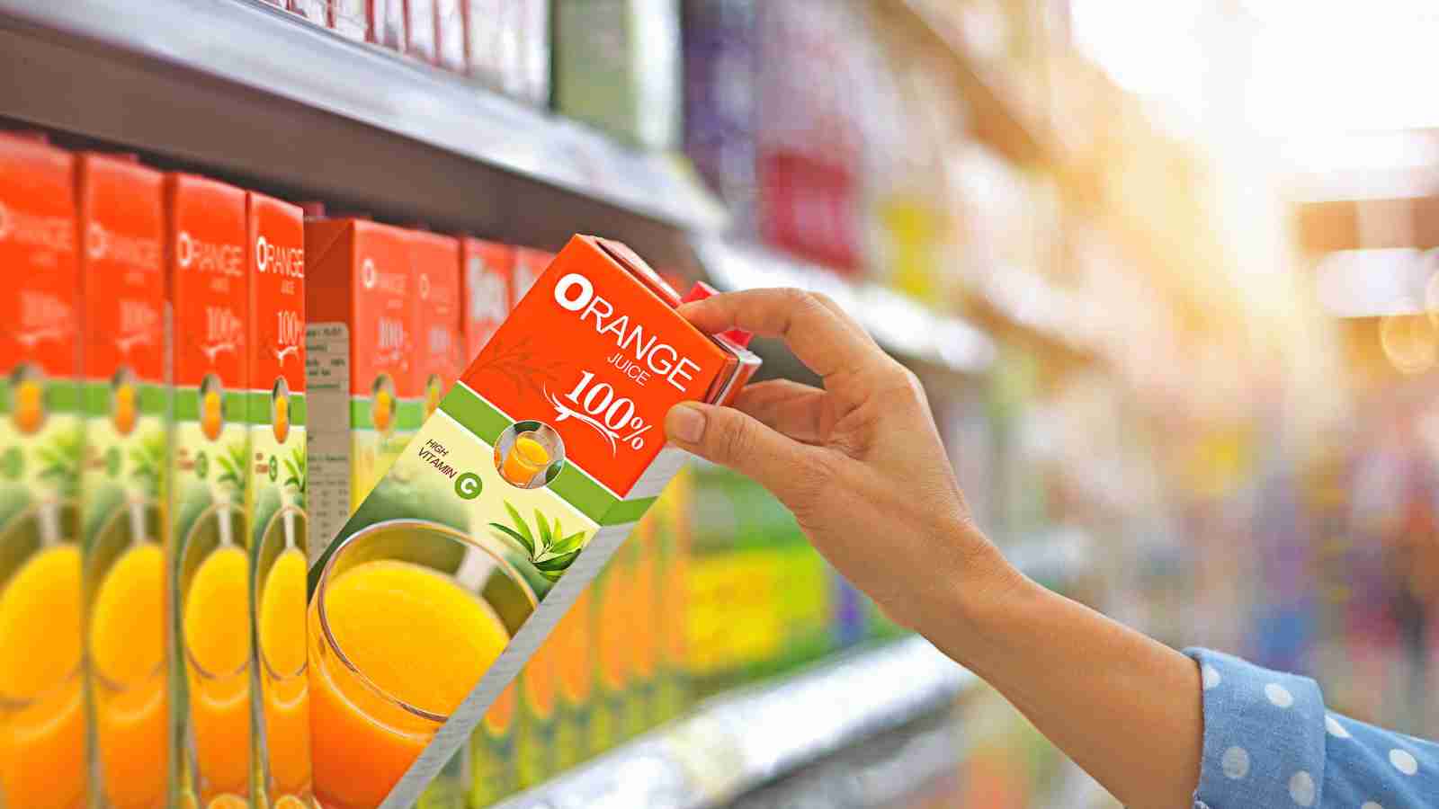 A hand reaches for a carton of 100% orange juice on a supermarket shelf.