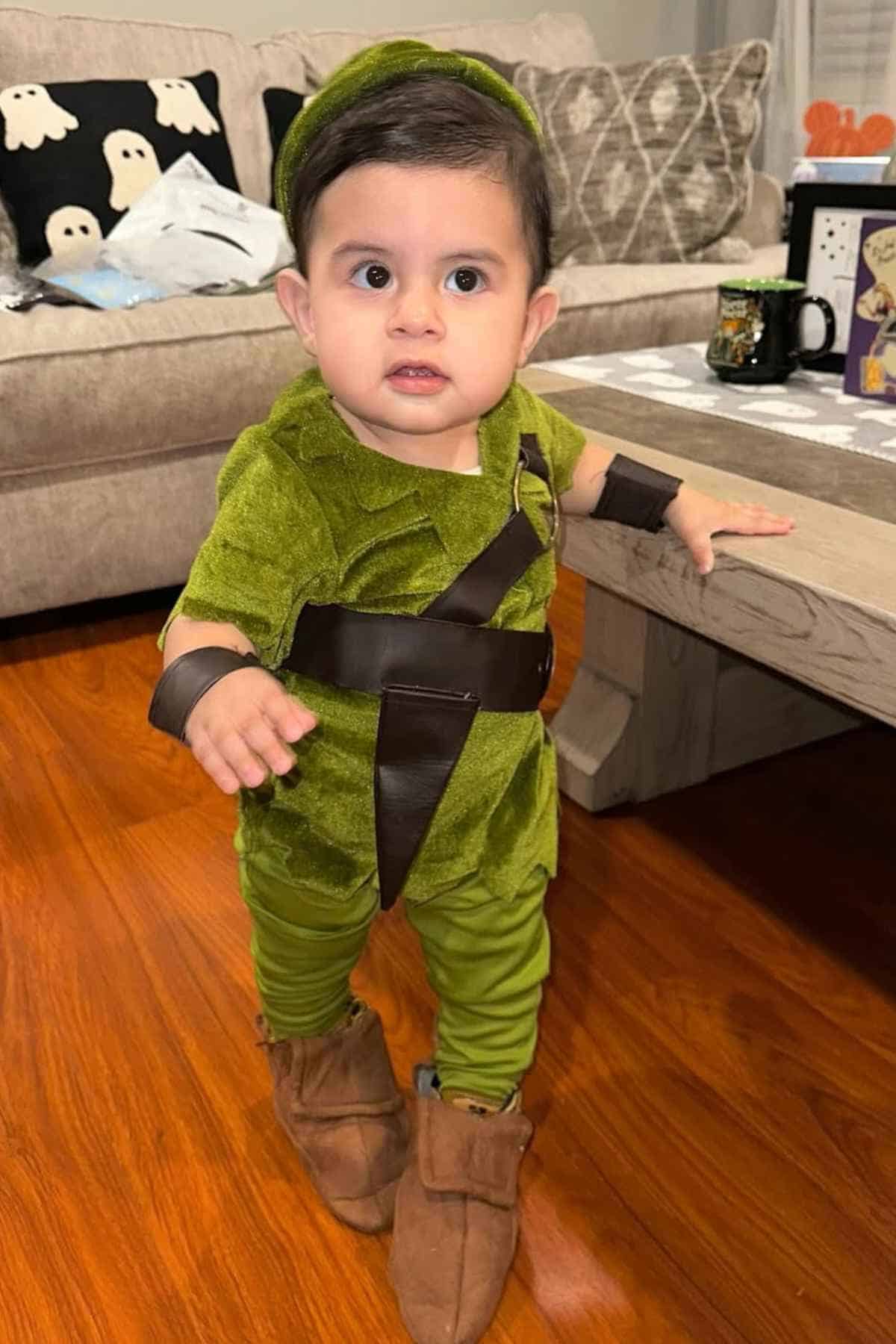 A toddler dressed in a green costume with brown accessories stands indoors on a wooden floor, holding onto a table for support.