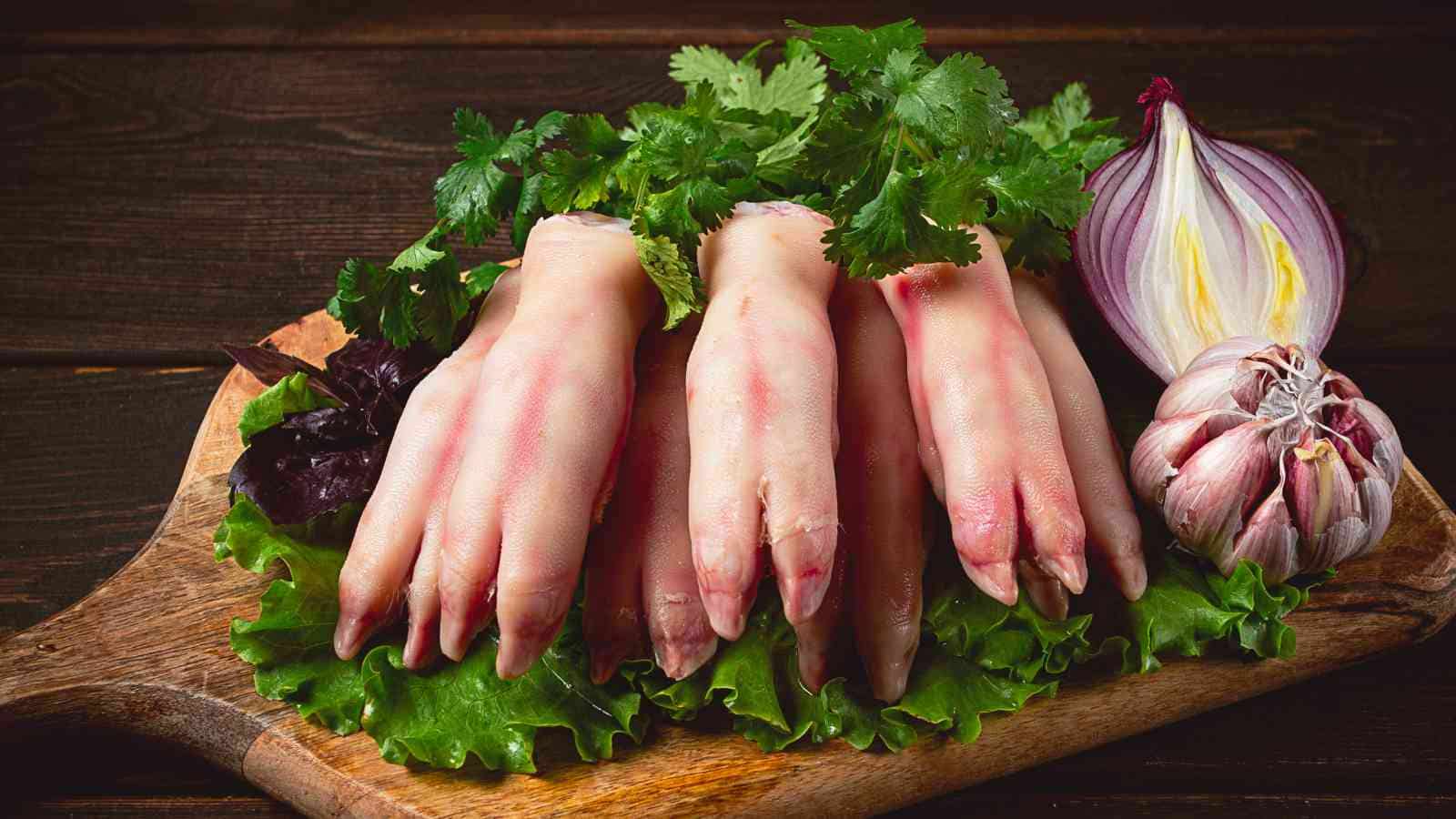 Several raw pig's feet are arranged on a wooden cutting board with lettuce, cilantro, a red onion, and a bulb of garlic.