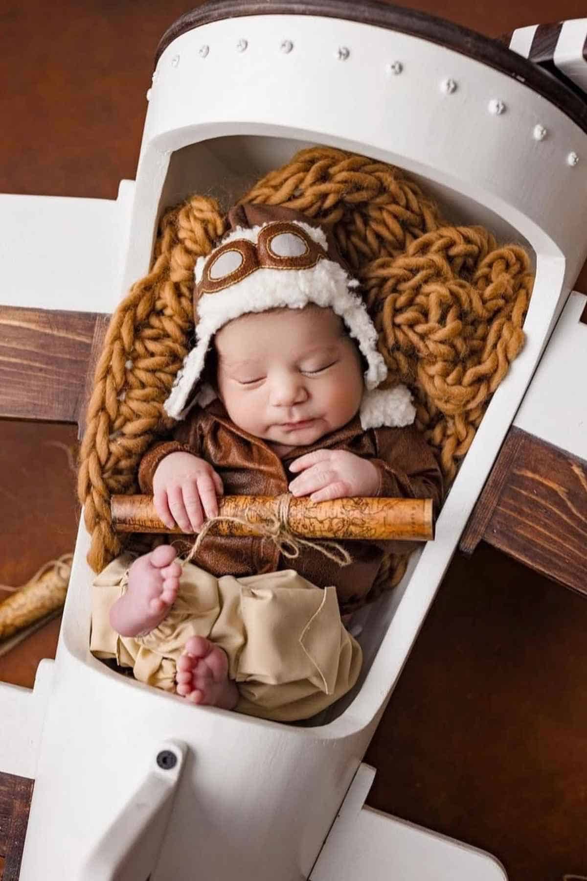 Baby lying on an aeroplane toy holding a rolled piece of paper, dressed as a pilot.