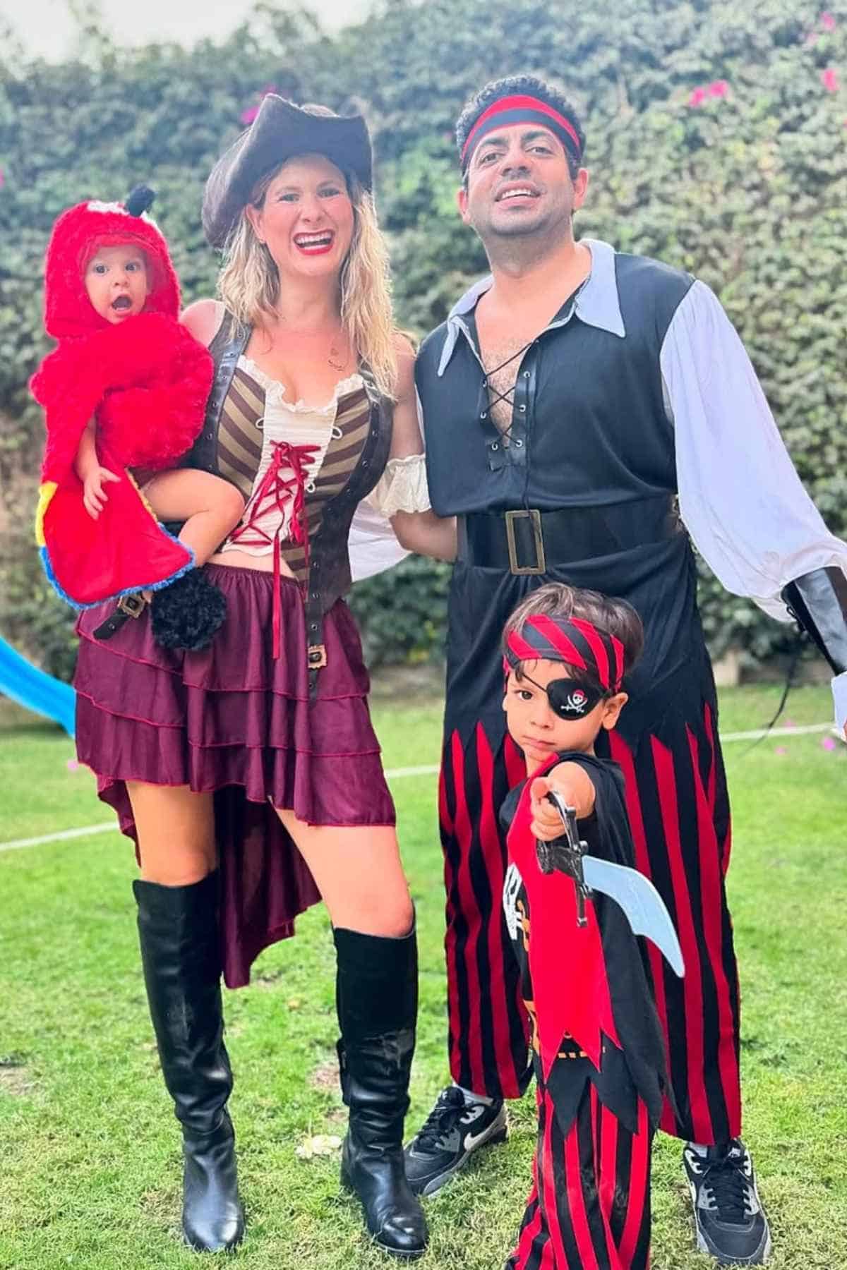 A family dressed in pirate and red monster costumes stands together outdoors on grass, with greenery in the background.