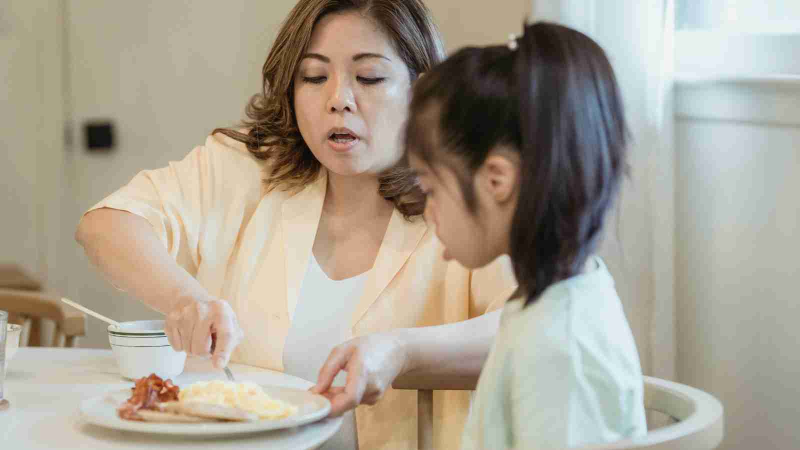 A woman serves a plate of breakfast food to a young girl at a table.