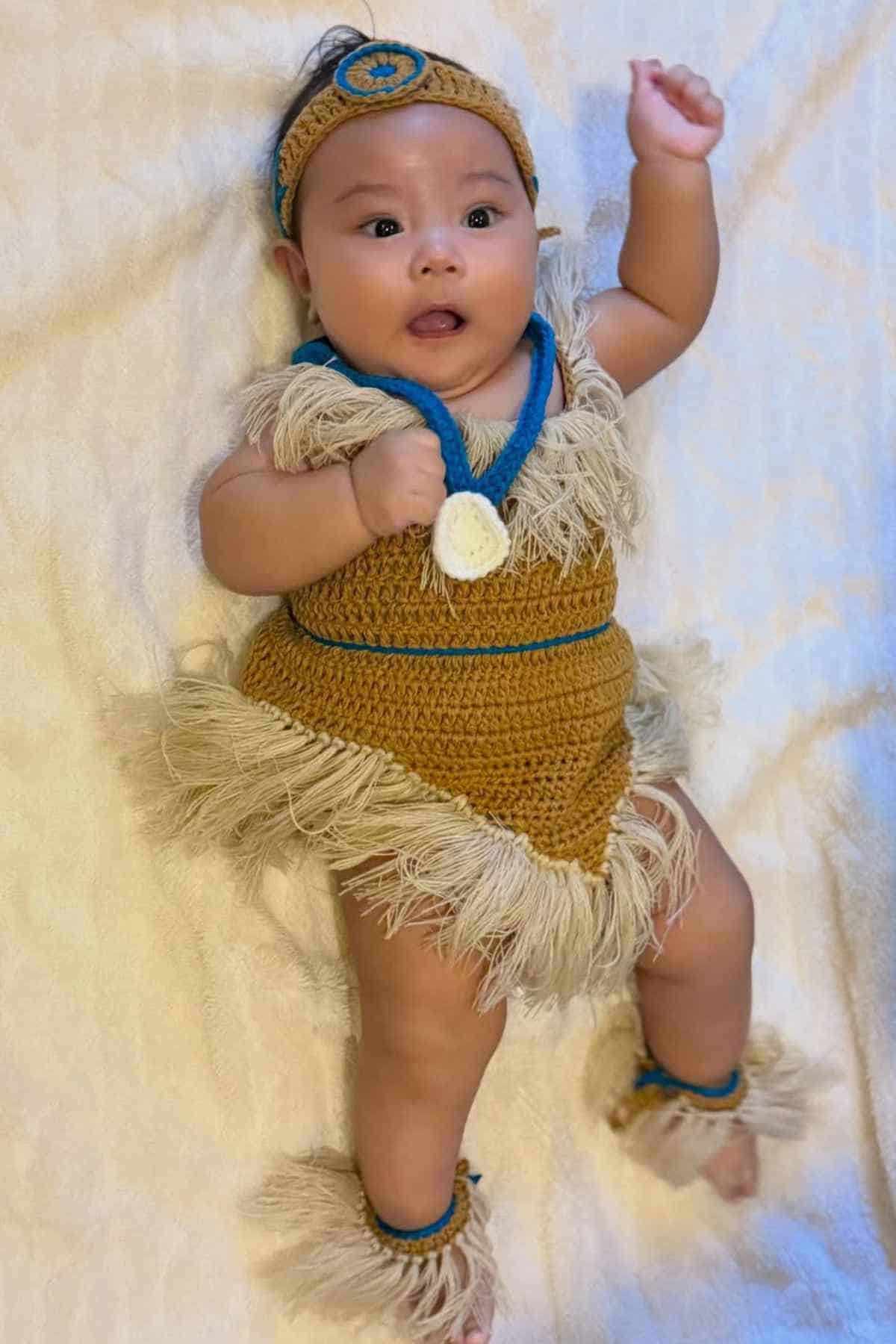 Baby lying on a cream blanket, wearing a crocheted tan and blue outfit with fringe, including a headband, dress, and anklets.