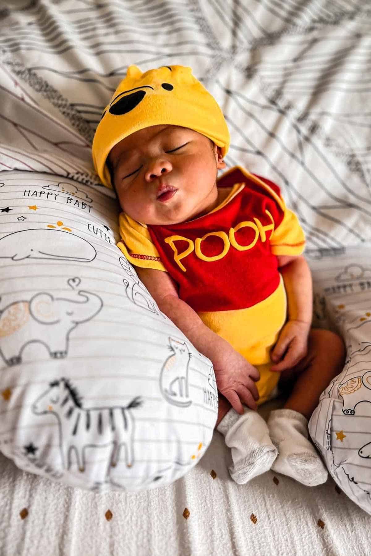 A baby lying a bed with some pillows, wearing a classic pooh costume paired with yellow hat.