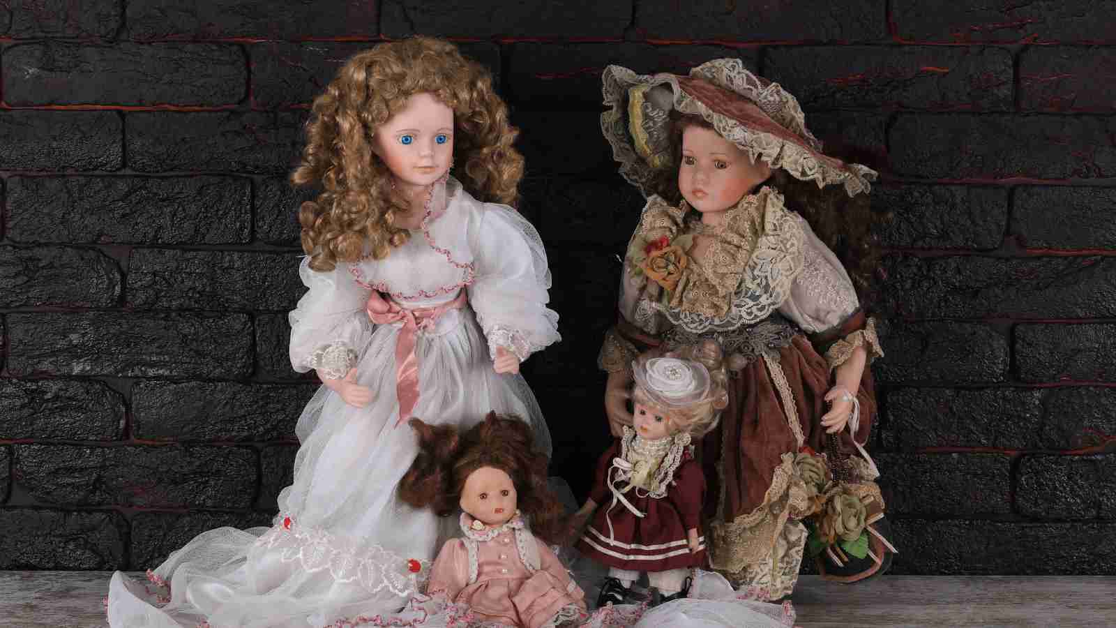 Three porcelain dolls in antique-style dresses are posed together against a dark brick wall.