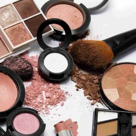 Assorted makeup products including blush, eyeshadow palettes, brushes, face powder, and a broken lipstick on a white surface.