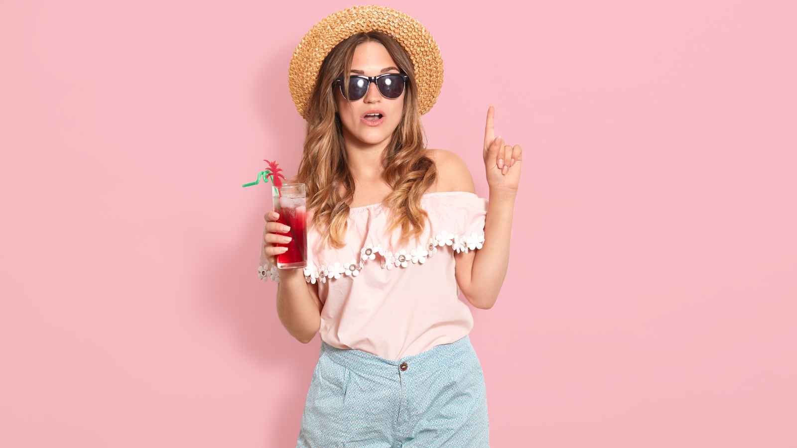 Woman wearing sunglasses and a straw hat holds a red drink in one hand and points upward with the other, standing against a pink background.