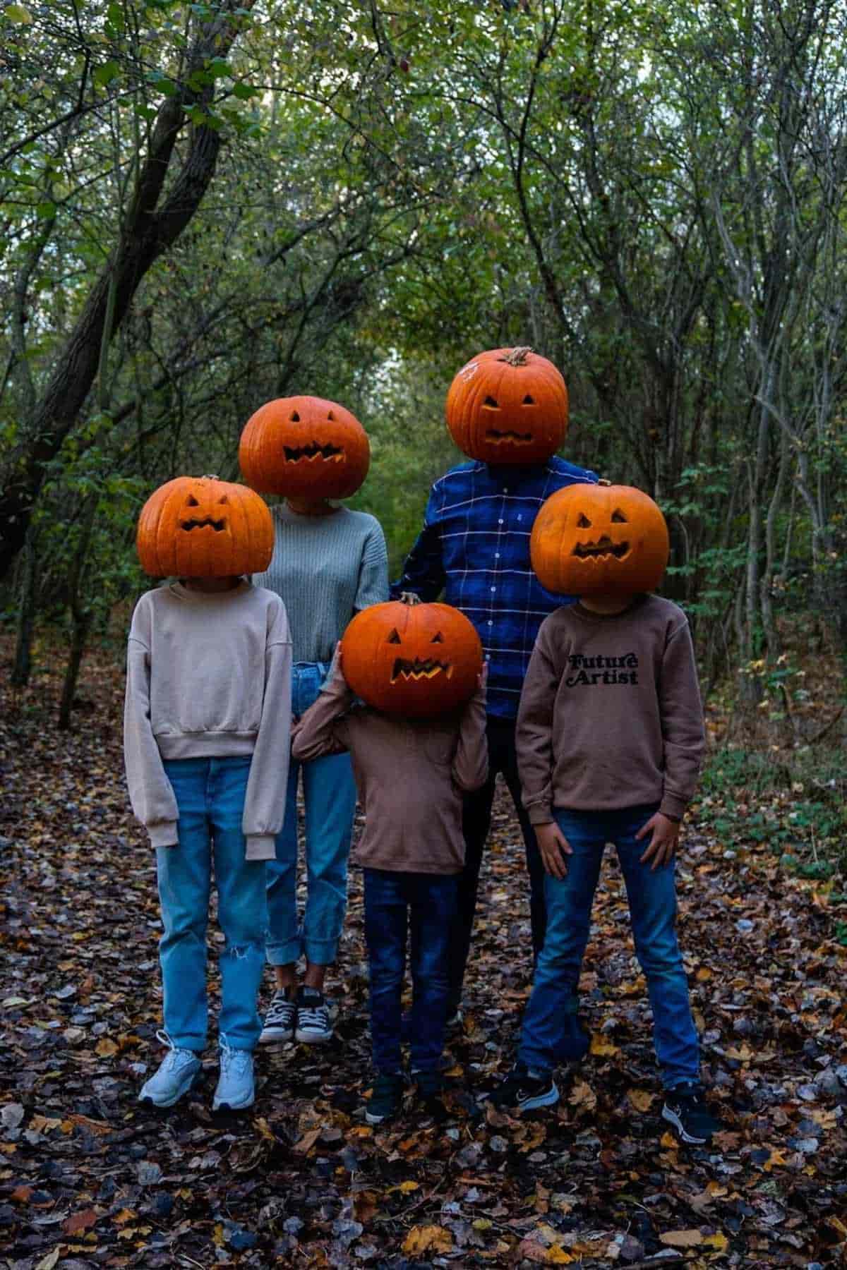 A family of five wearing pumpkin on their heads while standing straight in the middle of the woods.