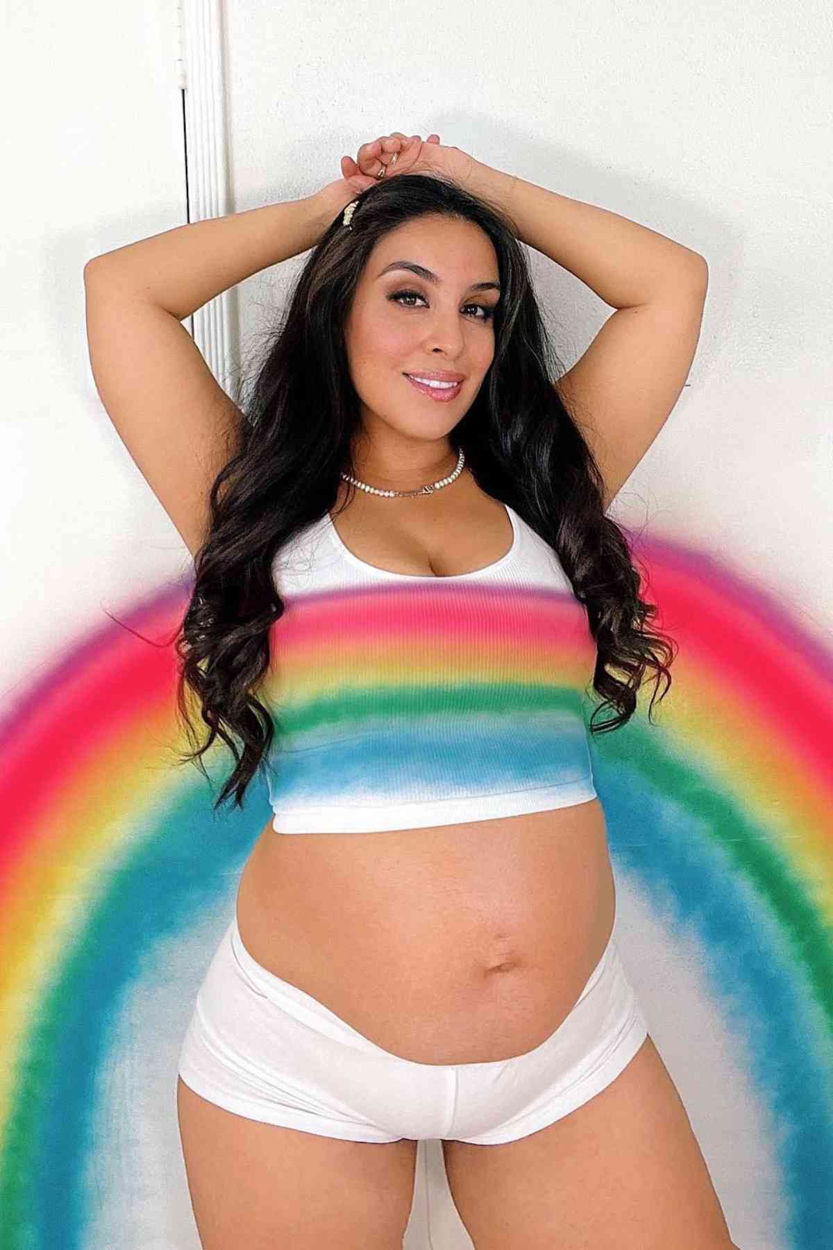 A pregnant woman stands against a white wall with a painted rainbow, wearing a white crop top and shorts, with her hands raised above her head.