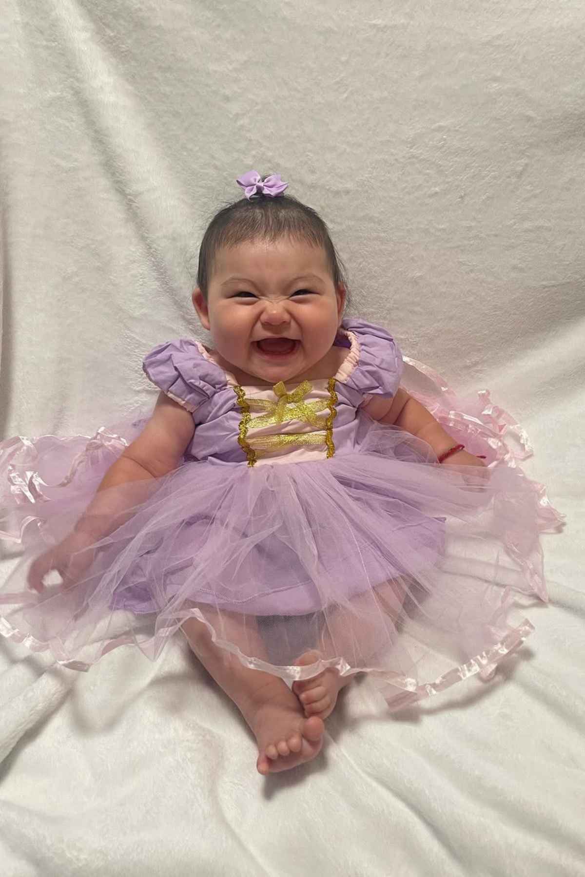 Smiling baby sitting on a white surface, wearing a purple and gold dress with a matching flower hair accessory.