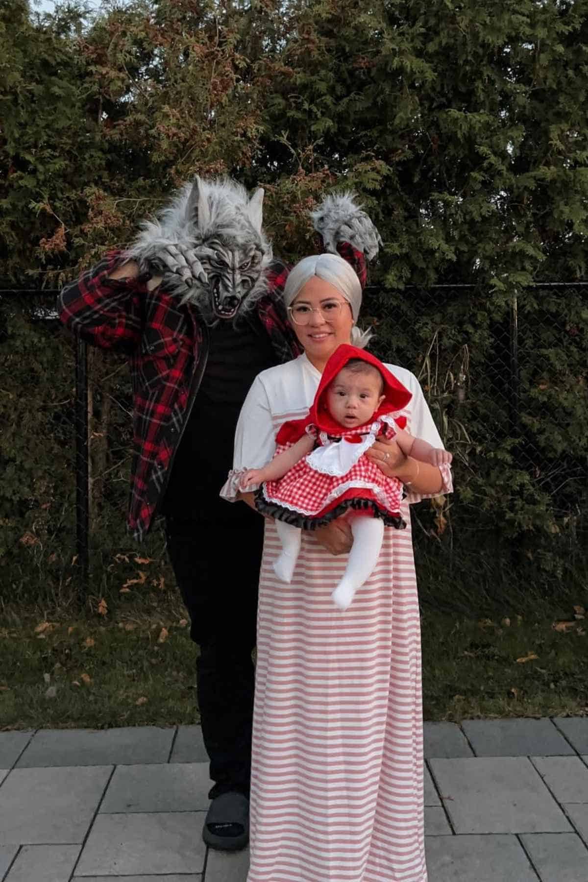 Three people in costumes stand outside: an adult in a wolf mask and plaid shirt, another adult dressed as a grandmother, and a baby dressed as Little Red Riding Hood.