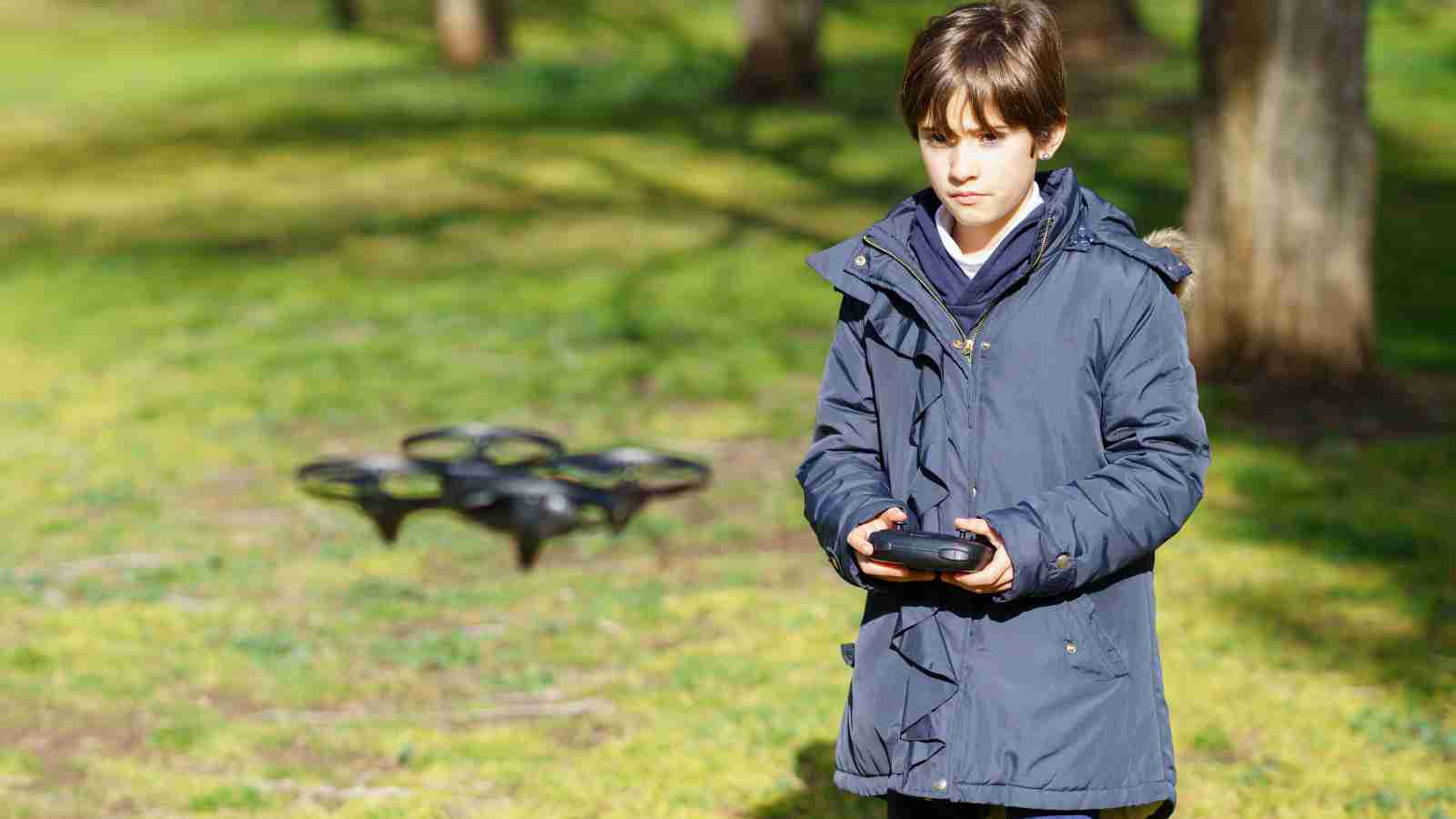A boy in a blue coat operates a remote control outdoors, with a drone on the grass in the background.
