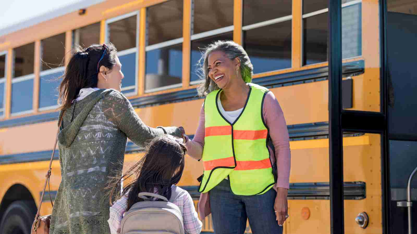 A school bus driver shakes hands with a young girl and her mother as they prepare to board the bus.