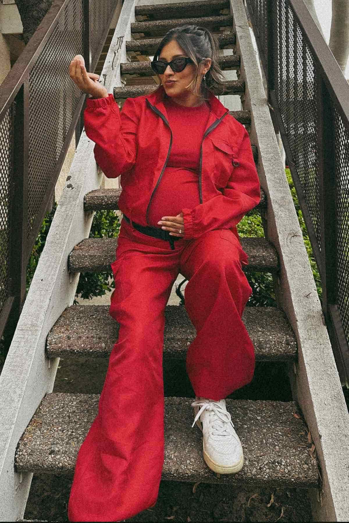 A pregnant woman in a red tracksuit and white sneakers sits on outdoor stairs, wearing sunglasses and holding one hand up.