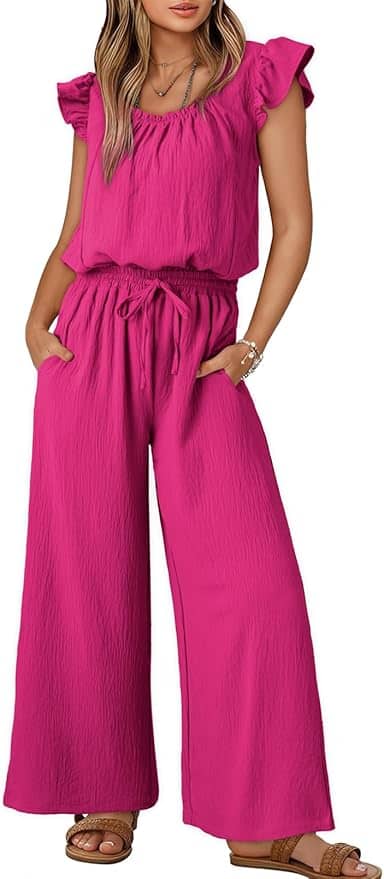 A woman wearing a bright pink, short-sleeve, ruffled jumpsuit with wide-leg pants and brown sandals, standing with her hands in her pockets.