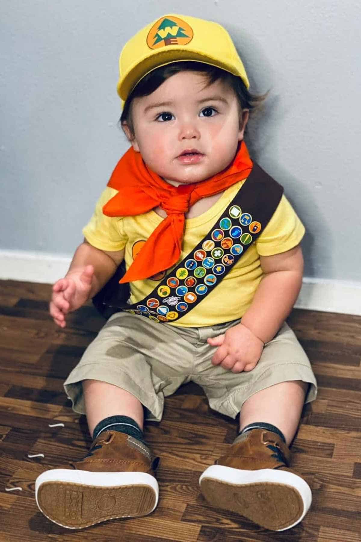 Young child sitting on a wooden floor, dressed in a yellow scout-themed outfit with a hat, orange neckerchief, and brown sash with colorful badges.